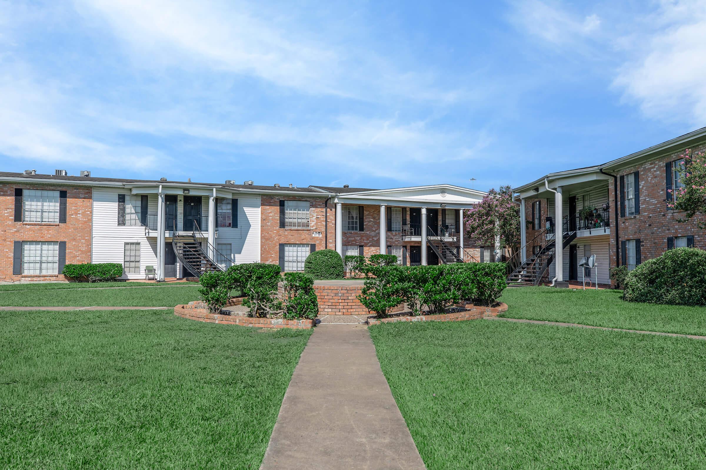 A well-maintained multi-unit apartment complex featuring two buildings with brick exteriors and green lawns. The central area has trimmed hedges and a pathway leading to entrances. Blue sky and light clouds create a bright atmosphere.