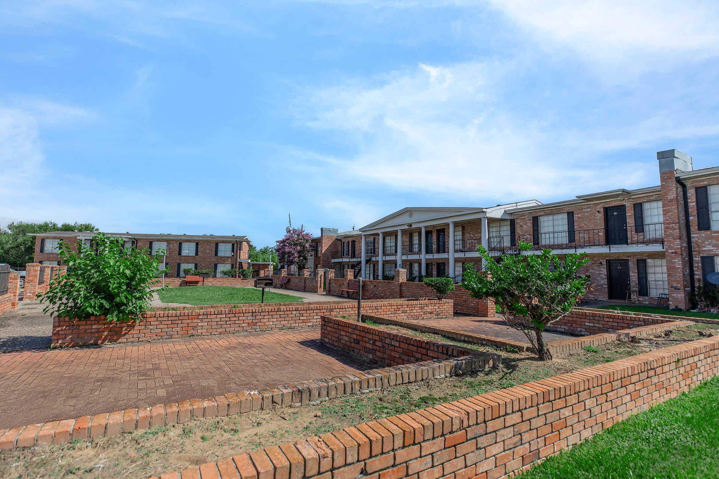 A residential area featuring brick buildings with multiple units, surrounded by well-maintained green spaces and landscaping. The sky is partly cloudy, and there are trees and shrubs in the foreground, enhancing the aesthetic appeal of the environment.