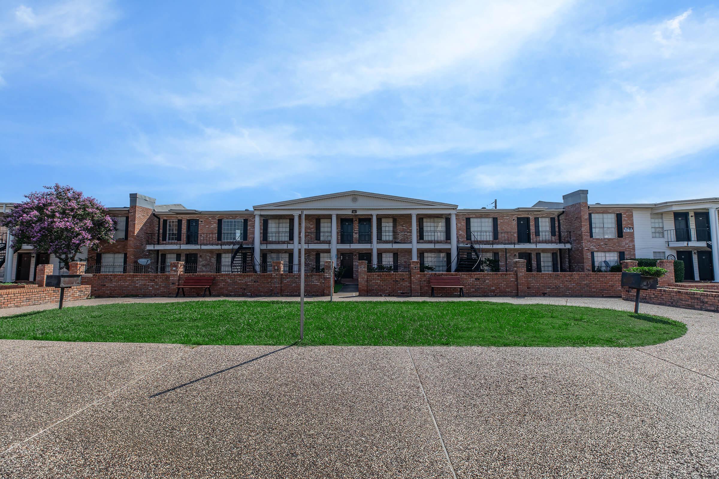 A view of a two-story brick apartment building with a central entrance, surrounded by well-maintained grass. Benches are placed along the path, and a flowering tree adds a touch of nature to the scene. The sky is bright with a few clouds, creating a pleasant atmosphere.