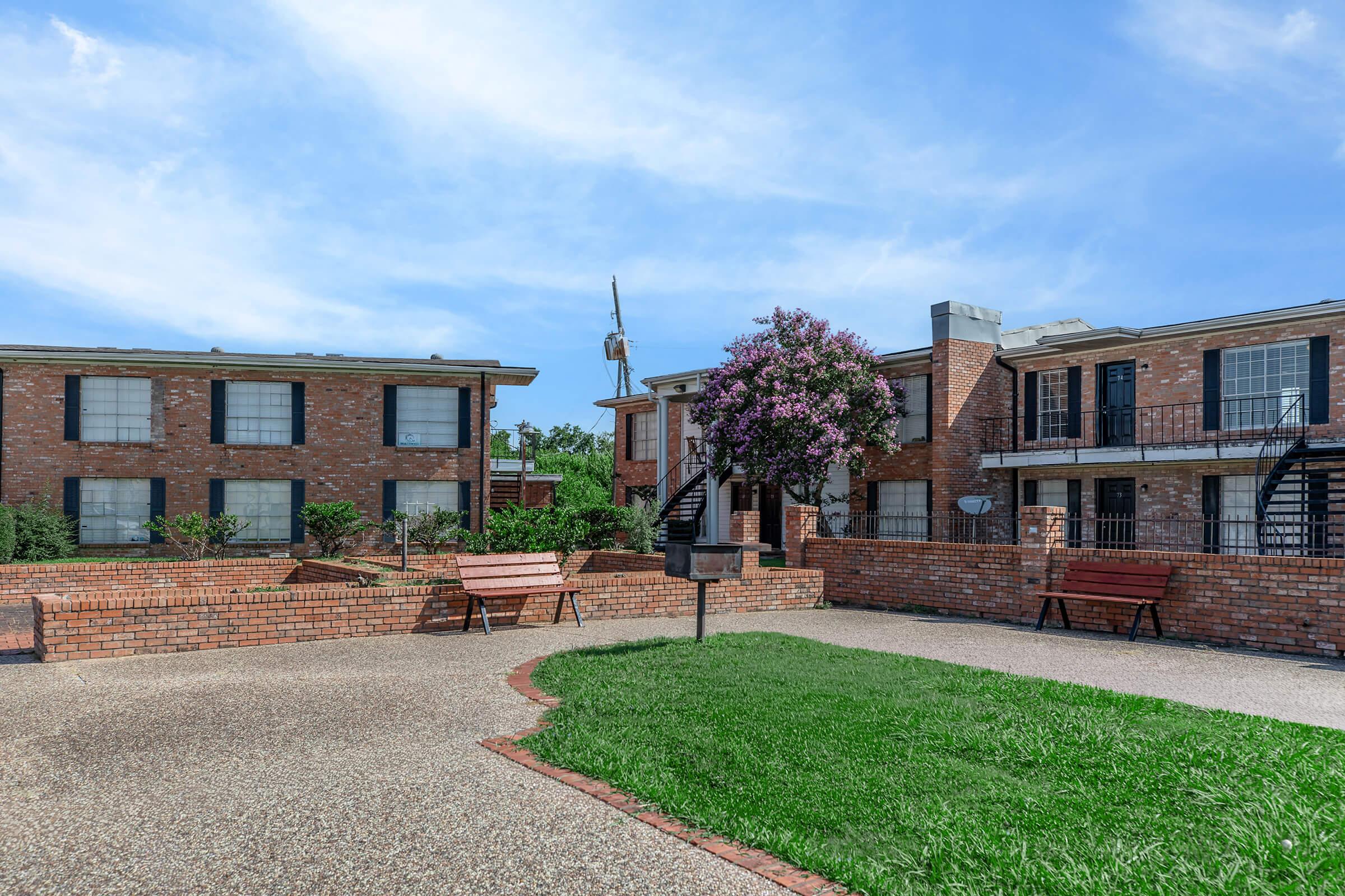 A view of a residential complex featuring brick buildings, grassy areas, and benches in a courtyard. The scene includes a flowering tree and a grill area, with blue skies in the background.