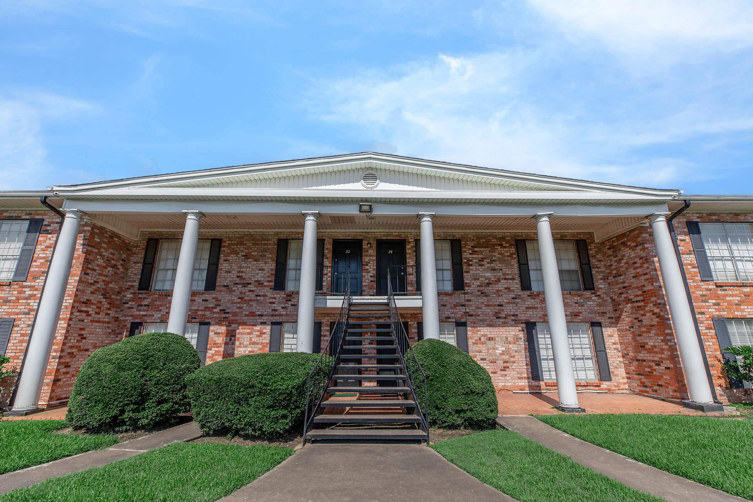 Exterior view of a two-story brick building featuring a central staircase flanked by manicured bushes, with large windows on either side. The building has white columns supporting a porch, set against a bright blue sky. The pathway leading to the entrance is lined with well-maintained grass.