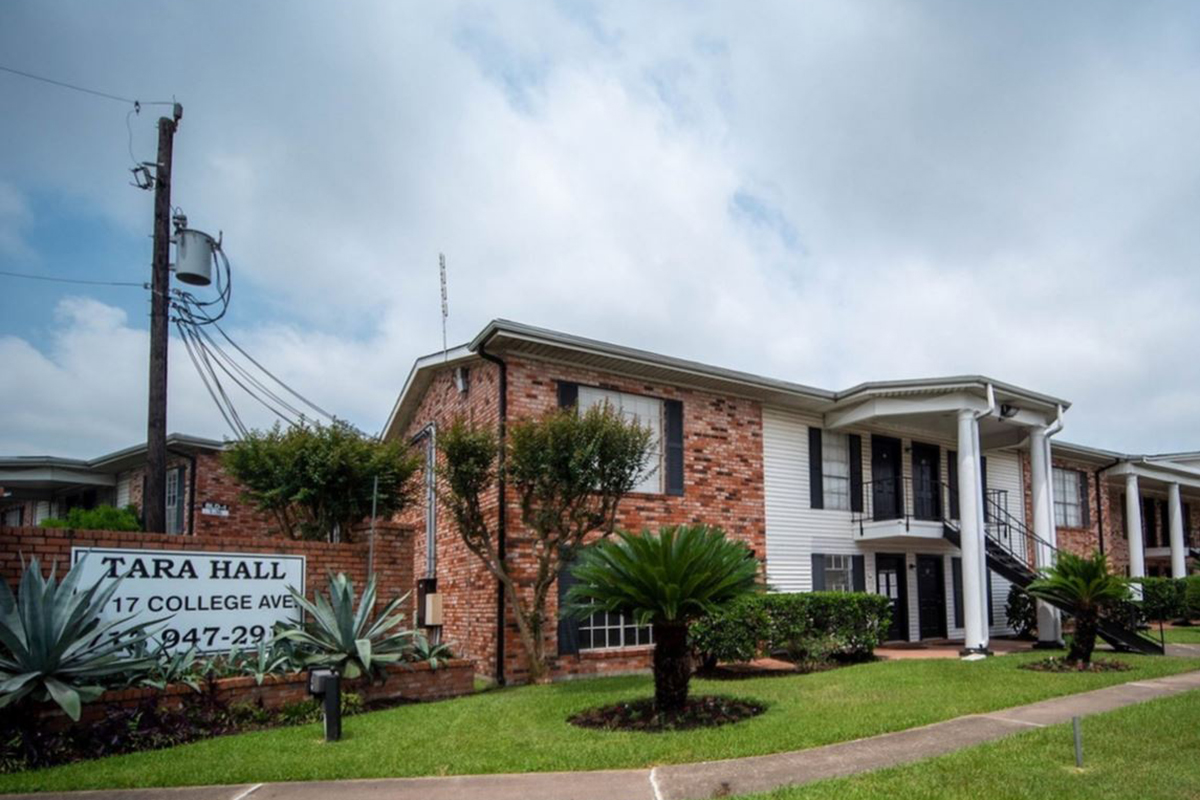 Brick apartment building with a sign reading "TARA HALL, 17 COLLEGE AVE" in front. The building has two stories, with a staircase leading to the second floor. There are neatly landscaped bushes and plants in the foreground, with power lines visible in the background under a partly cloudy sky.