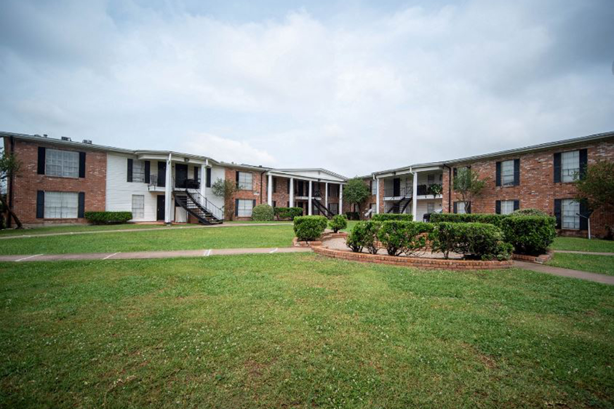 A view of an apartment complex featuring two brick buildings surrounded by green lawns and shrubs. The buildings have multiple balconies, and there are walkways leading through the grassy area. The sky is cloudy, giving a serene ambiance to the setting.