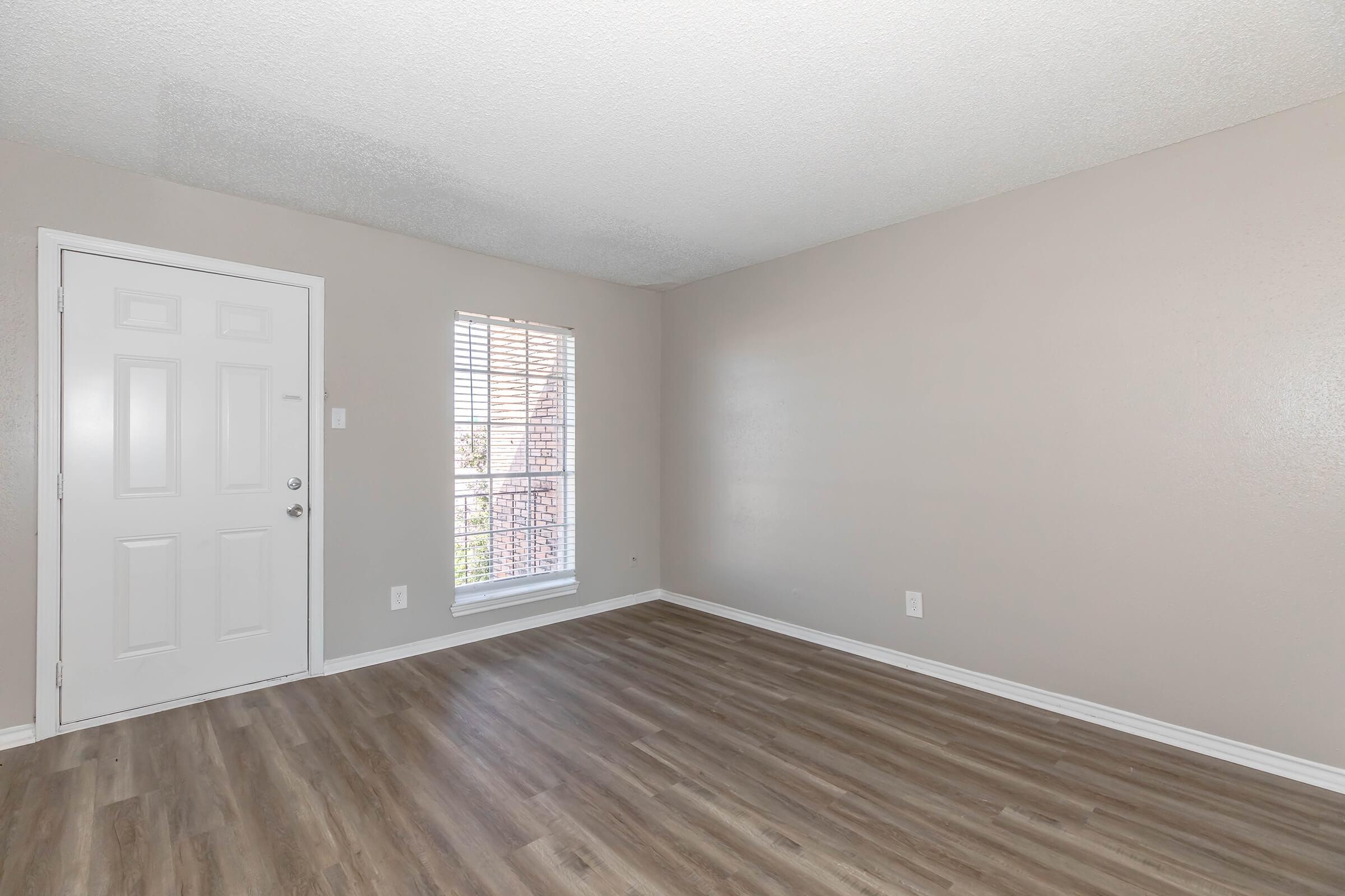 Empty room with light brown walls and laminate flooring. A door is visible on the left, and a window with blinds is on the right, letting in natural light. The space is unfurnished, creating a clean and simple aesthetic.