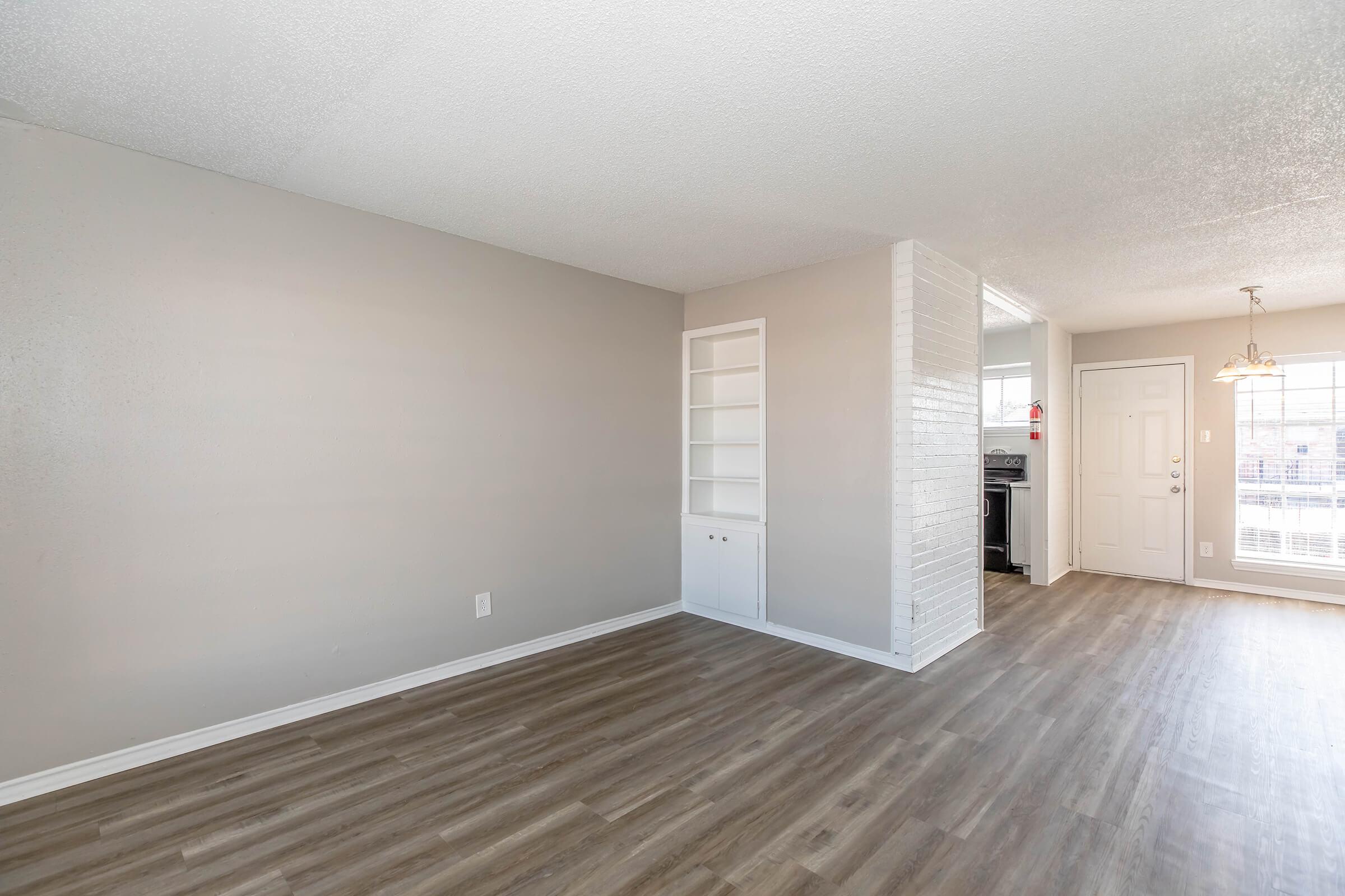 Spacious, empty living area with light gray walls and vinyl flooring. There is a built-in shelving unit on the right side. The room features a door leading outside and a window allowing natural light. A glimpse of the kitchen can be seen through an opening in the wall.