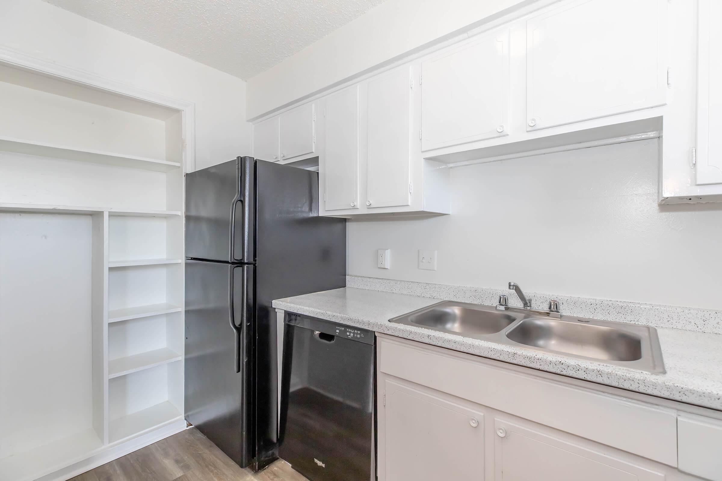 A modern kitchen featuring white cabinetry, a stainless steel double sink, and a granite countertop. Appliances include a black refrigerator and a black dishwasher. There is also a pantry or shelving unit next to the refrigerator, providing additional storage space. The flooring is a light wood laminate.