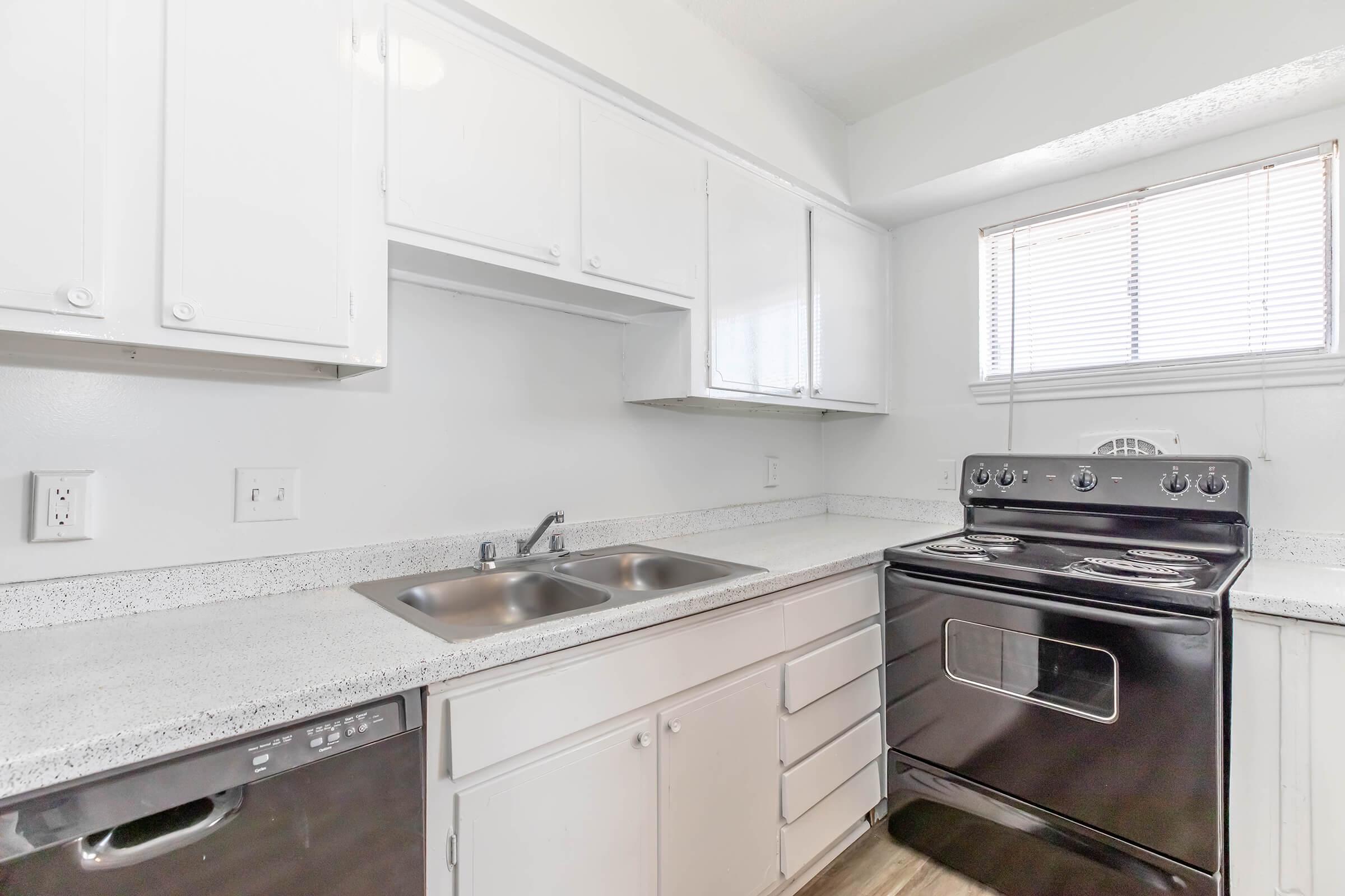 Modern kitchen featuring white cabinetry, a dual sink with a faucet, a black stovetop oven, and a dishwasher. Natural light enters through a window with blinds. The countertops are light-colored with a speckled design, providing a clean and contemporary look.