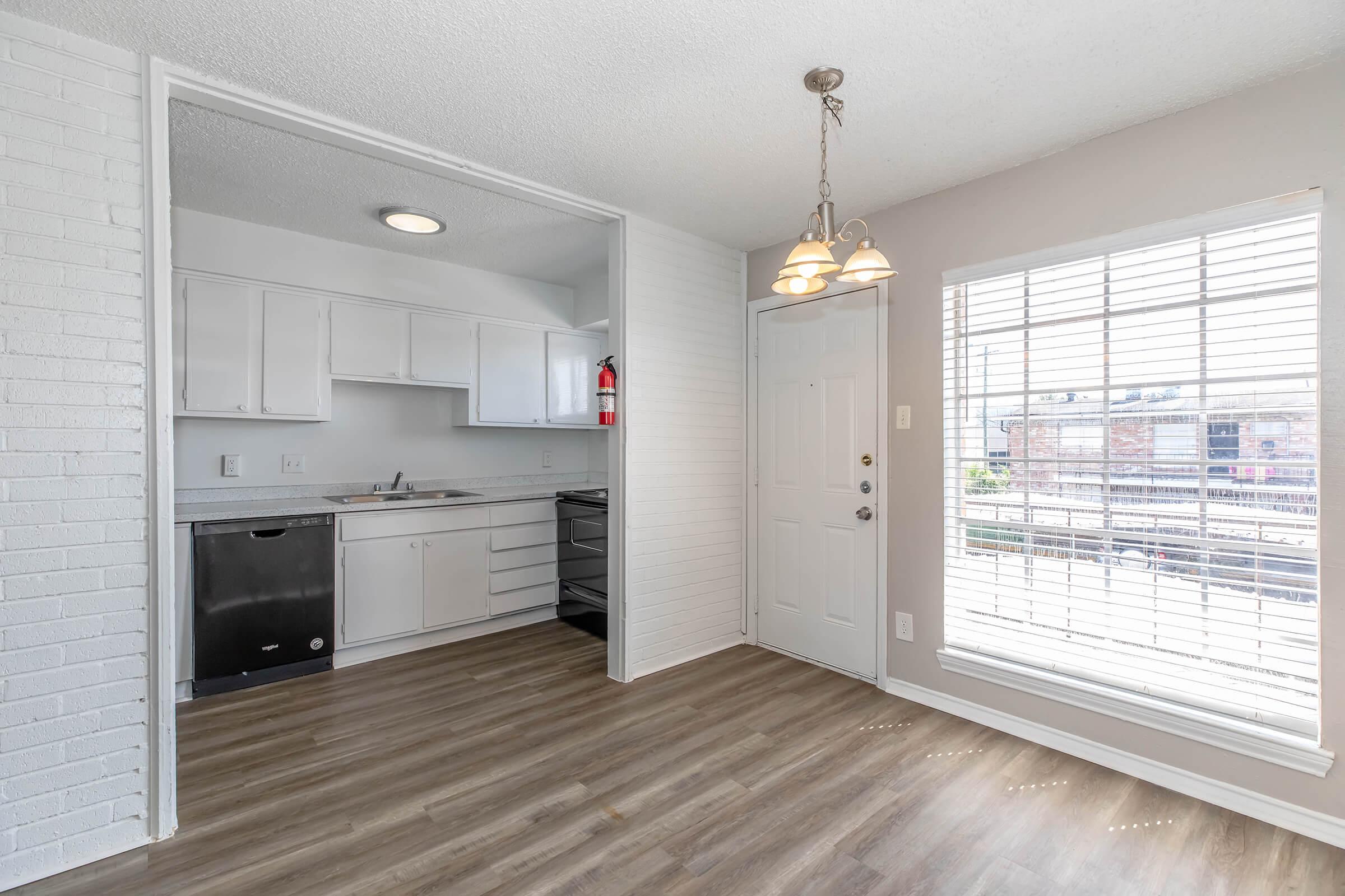 A modern kitchen and dining area featuring white cabinetry, a dark dishwasher, and a stove. There's a window with blinds allowing natural light, and a doorway leading outside. The flooring is a light wood finish, creating a spacious and inviting atmosphere. A light fixture hangs above the dining area.