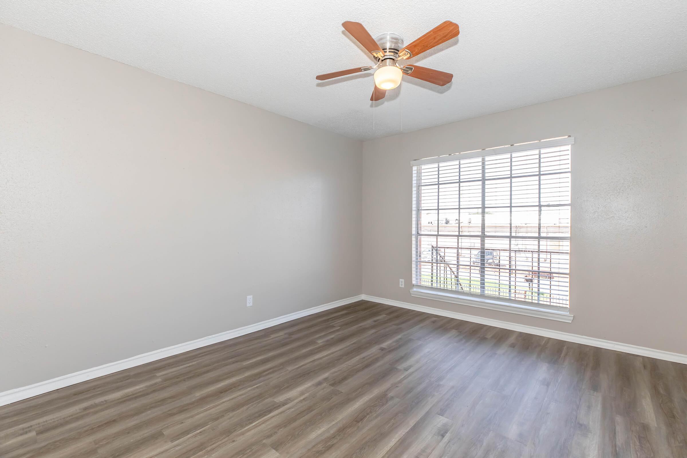 A spacious, empty room with light brown walls and a ceiling fan. The floor is made of laminate wood, and there is a window with white blinds allowing natural light to enter. The room has a clean, minimalistic design, perfect for various decor styles.