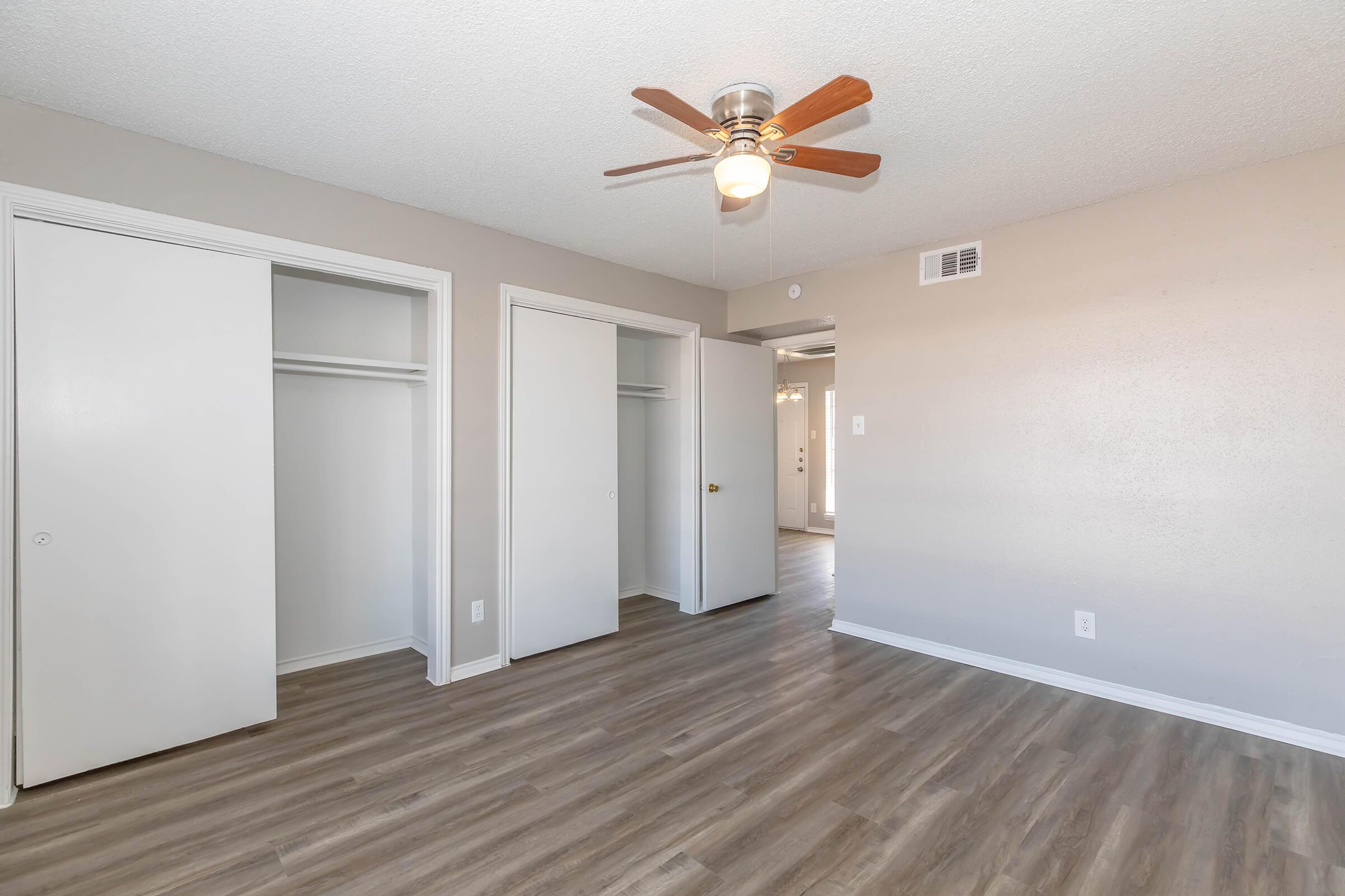 A spacious room with light-colored walls and vinyl flooring, featuring a ceiling fan with wooden blades. Two white closet doors are visible on one side, and the room is illuminated by natural light. A doorway leads to another area, suggesting an open layout.