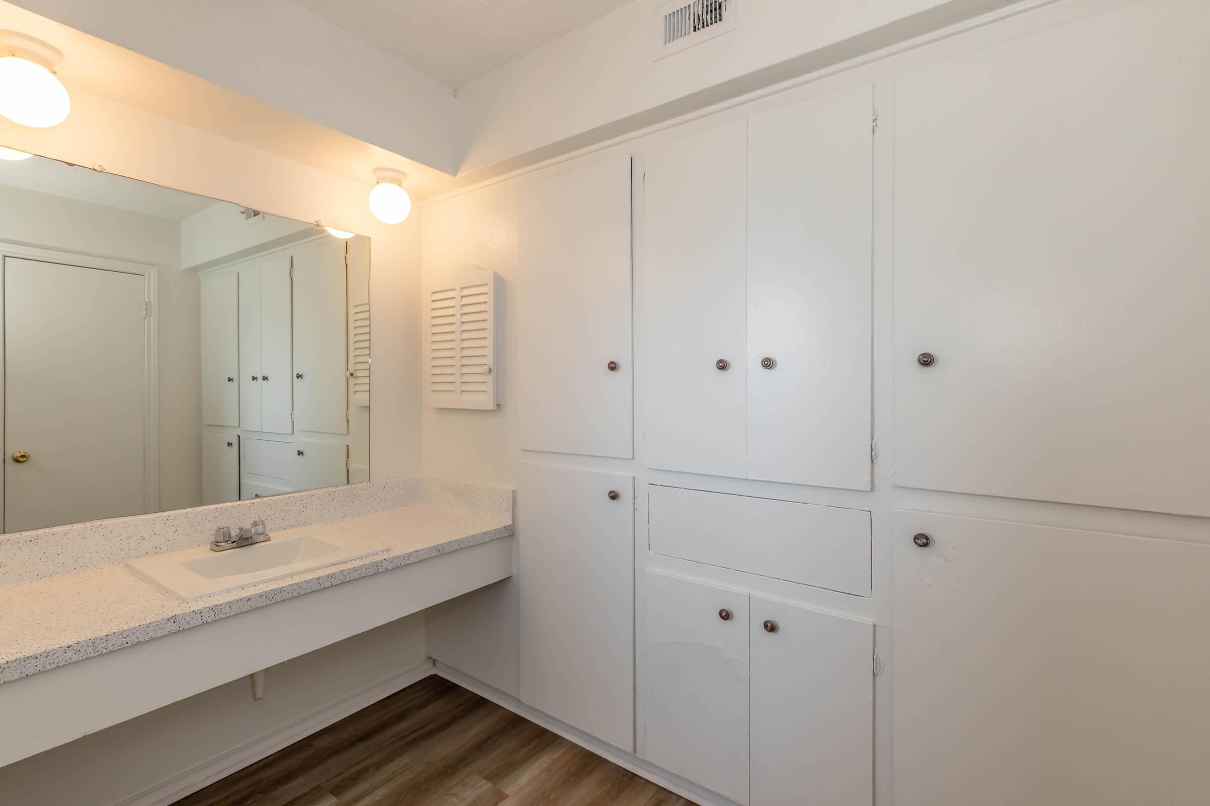A clean, minimalist bathroom featuring a countertop with a sink, white closed cabinets above and below, a large mirror with warm lighting, and a light-colored wood floor. The overall design is simple and functional, emphasizing a bright and spacious feel.