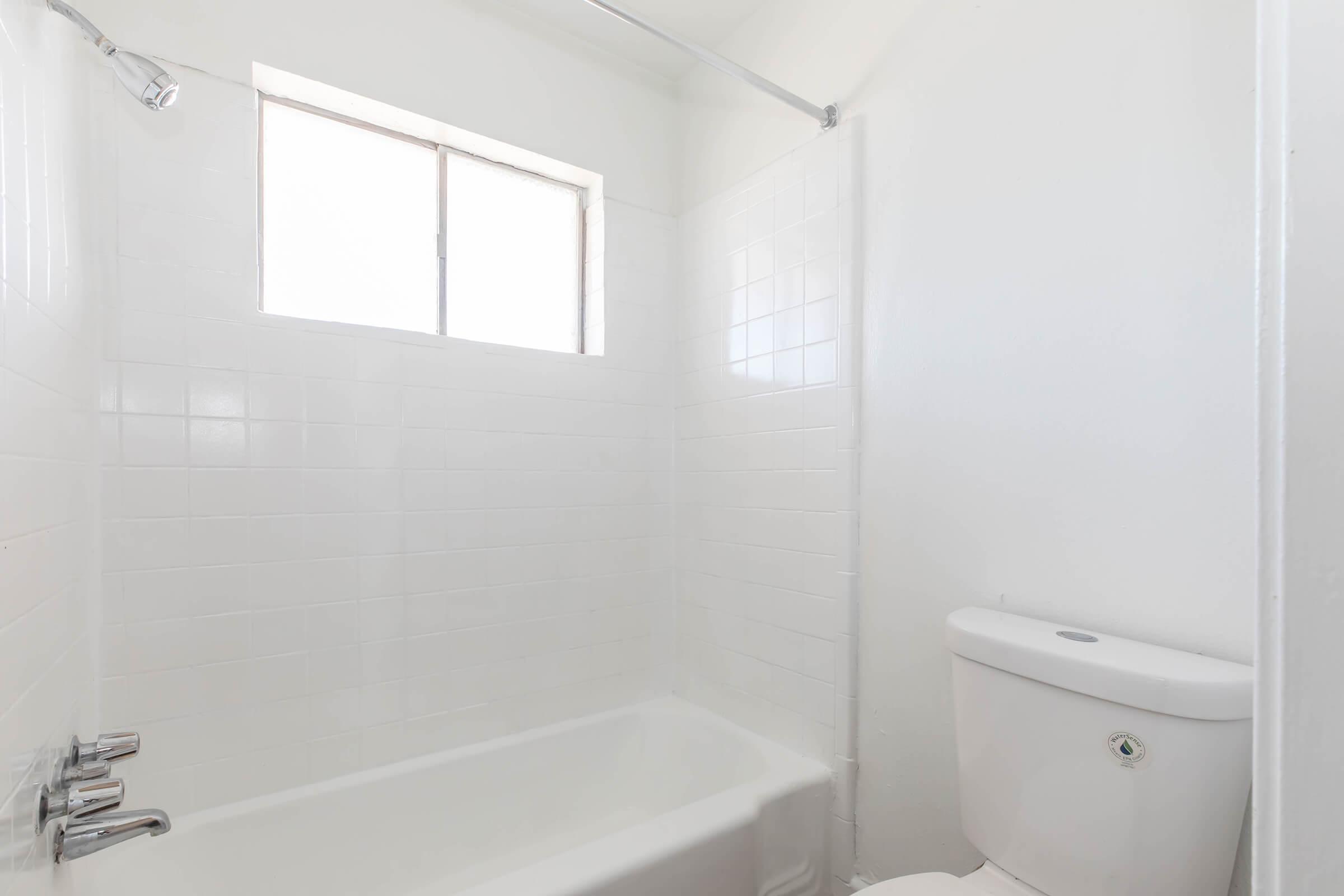 A clean and minimalistic bathroom featuring a bathtub with a showerhead, white tiled walls, and a toilet. A window allows natural light to brighten the space. The overall design is simple and uncluttered, emphasizing a fresh and modern look.