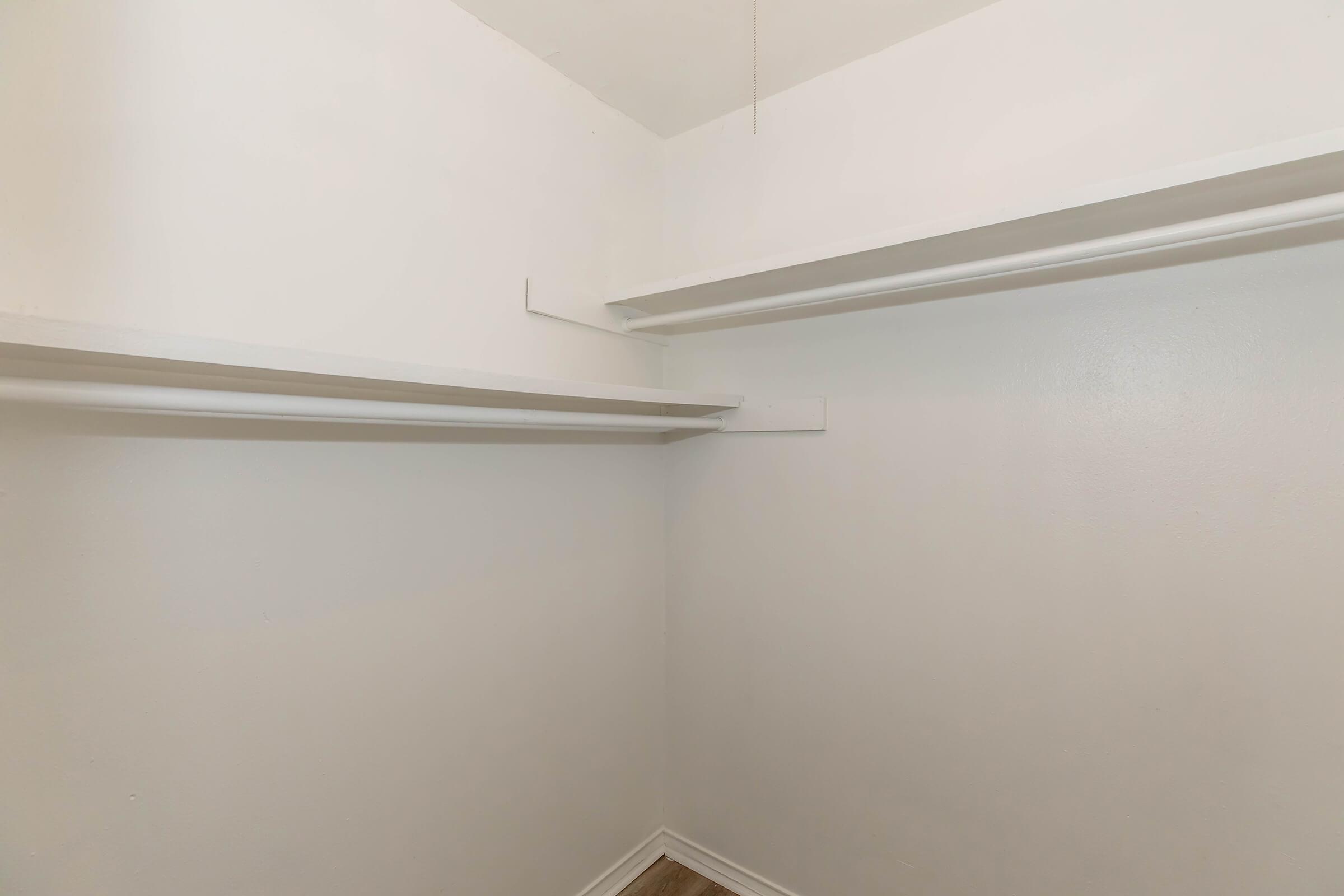 A tidy, white empty closet with two simple shelves on opposite walls. The walls are smooth with a clean finish, and the floor appears to have a light-colored, wooden look. The ceiling is plain with a light fixture overhead.