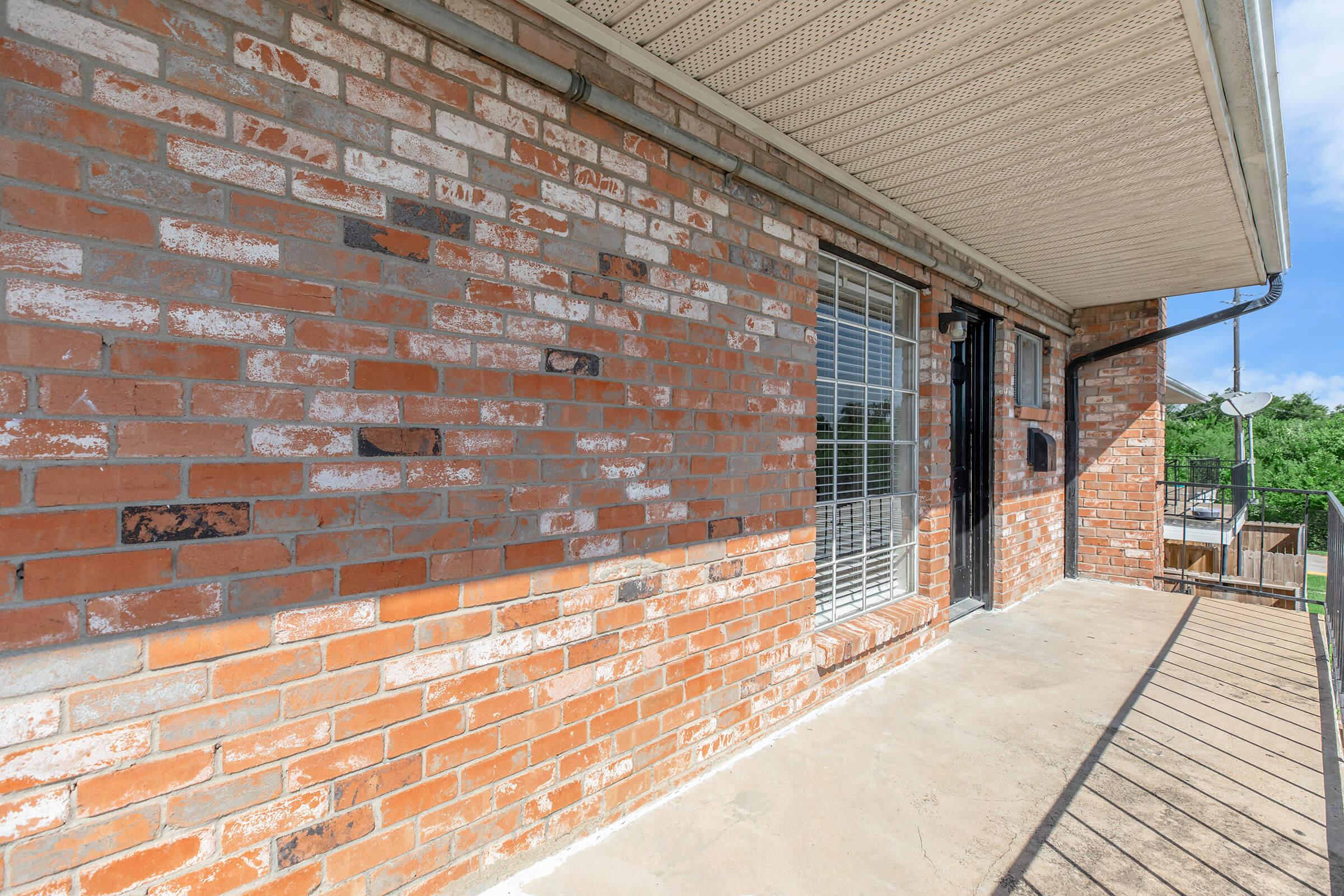 A close-up view of a brick exterior wall with a window and a door leading onto a small balcony. The bricks have a mix of red and gray tones, and the balcony features a railing. Natural light illuminates the space, highlighting the texture of the brickwork.