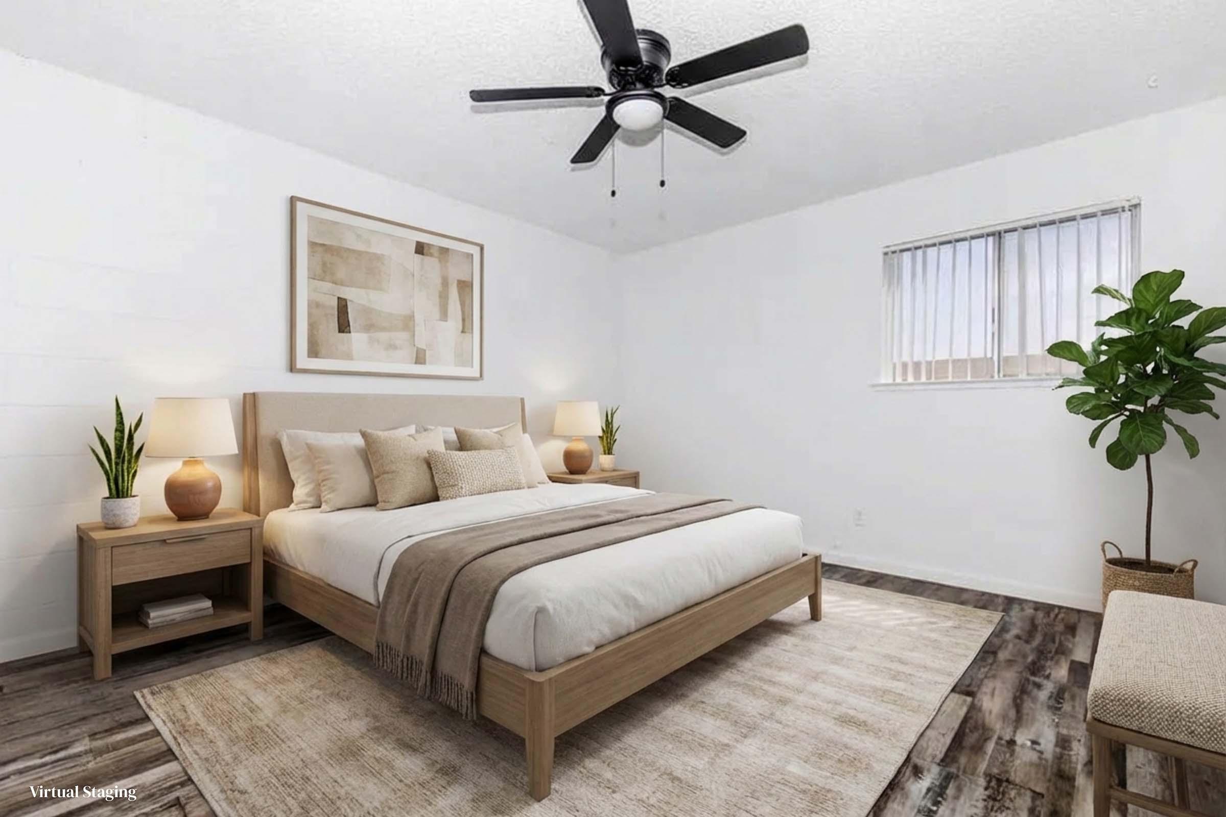 A modern bedroom featuring a light wood bed with neutral bedding, two bedside lamps, and a matching nightstand. A large area rug lies on the floor, and a ceiling fan is visible. A window with blinds allows natural light, and a potted plant adds a touch of greenery. Abstract wall art complements the minimalist decor.