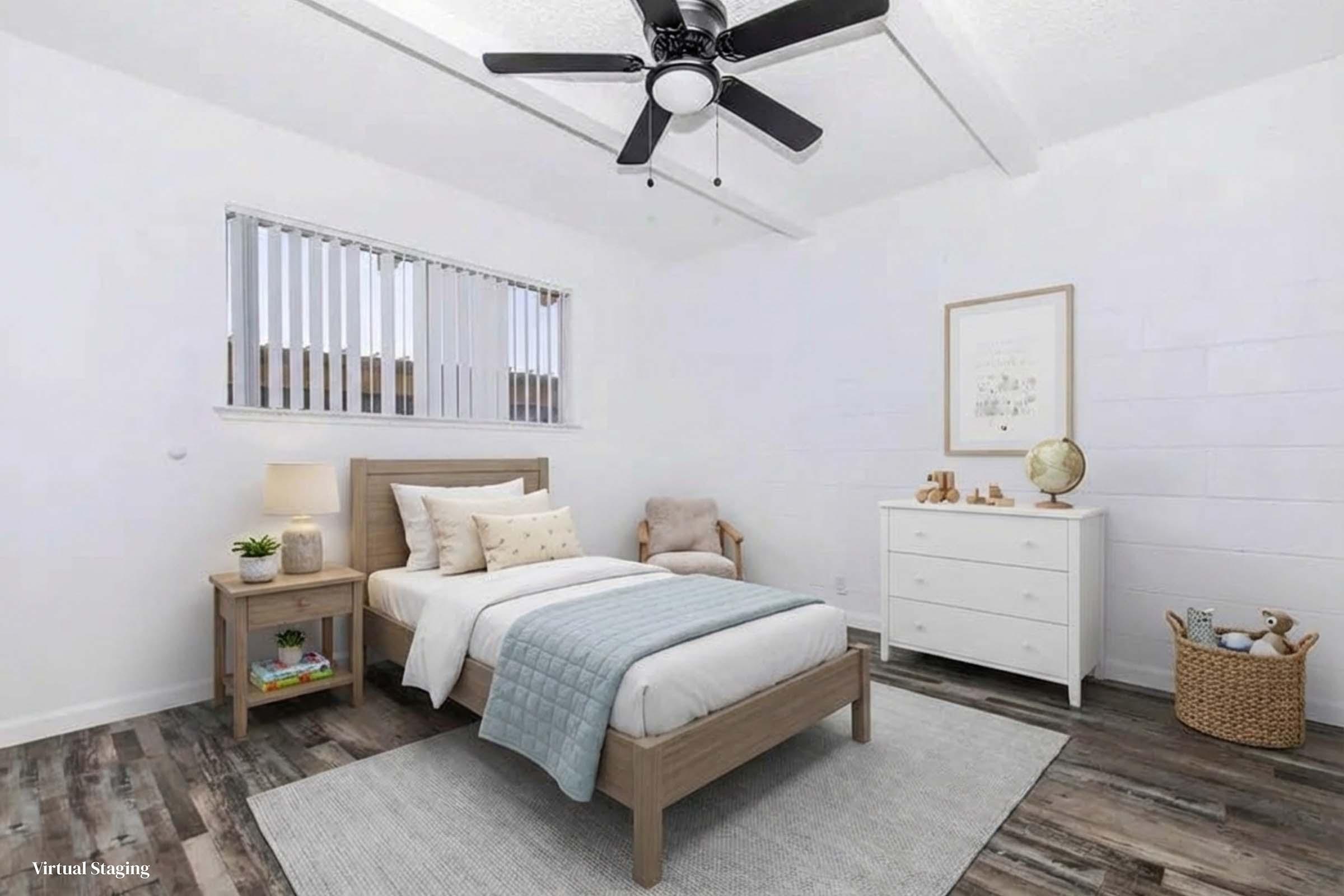 A bright and airy bedroom featuring a single bed with a light blue throw blanket, a nightstand with a lamp, a cozy armchair, a white dresser, and a wall-mounted fan. Natural light enters through horizontal blinds, and a soft area rug lies on the floor, creating a serene atmosphere.