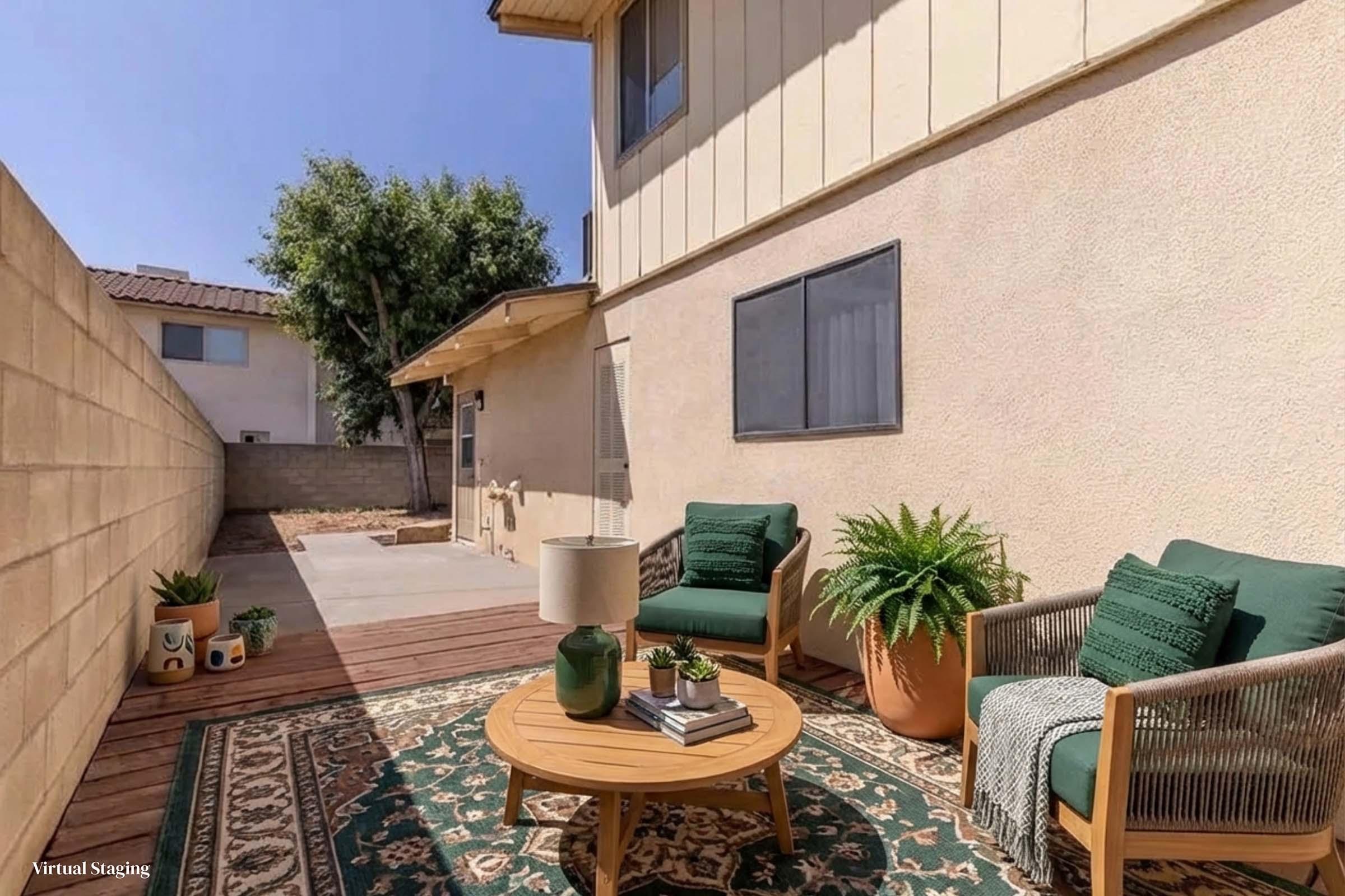 A cozy outdoor space featuring a wooden deck with a round coffee table at the center. Two green chairs are arranged around the table, accompanied by a potted plant and a lamp. The walls are beige, and there are trees visible in the background, creating a relaxing atmosphere.