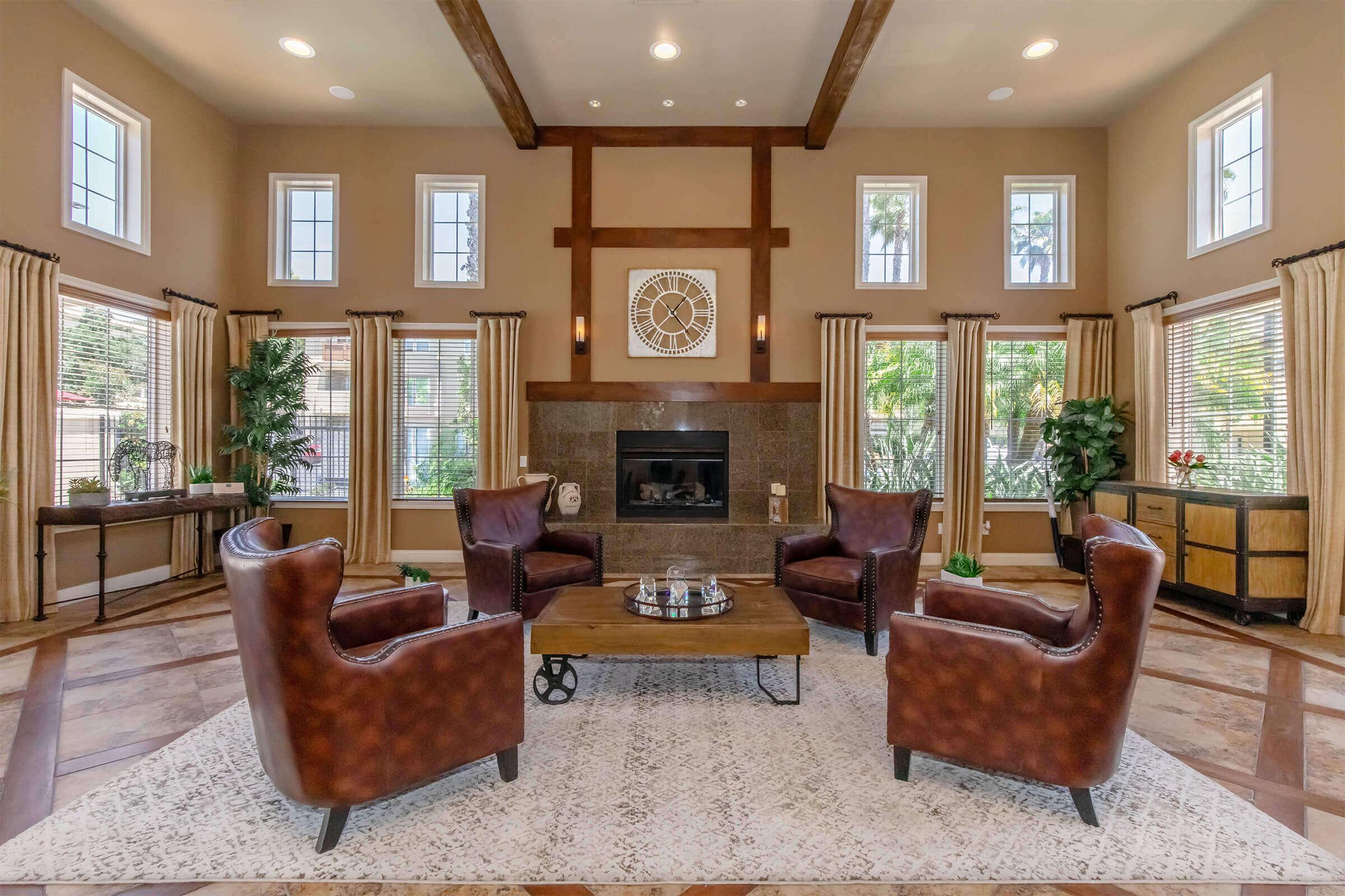 A spacious living room with high ceilings, featuring two brown leather armchairs, a wooden coffee table with decorative items, and a dark stone fireplace. Large windows allow natural light to illuminate the space. Elegant curtains frame the windows, and potted plants add a touch of greenery.