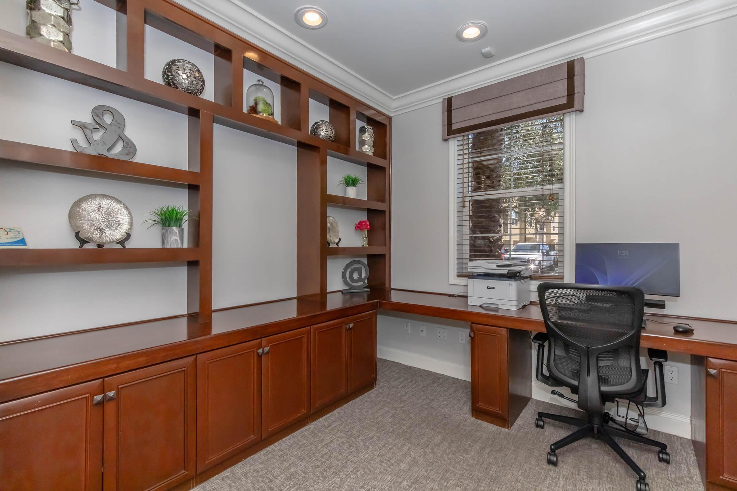 A modern home office featuring a wooden desk with a computer and printer. The wall is adorned with open shelves displaying decorative items, including sculptures and plants. Natural light enters through a window covered with a brown Roman shade, and the carpeted floor adds to the cozy atmosphere.