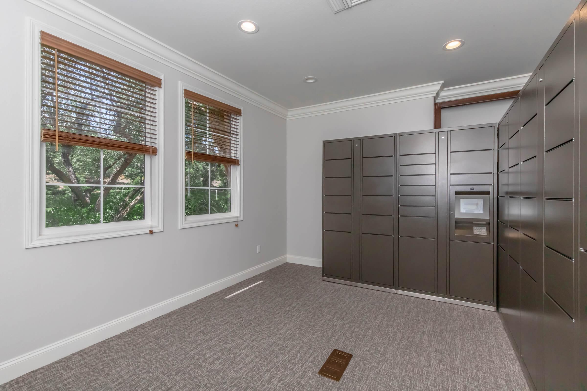 A modern indoor space featuring a light gray wall and a carpeted floor. There are two windows with wooden blinds allowing natural light, and a large storage locker system against one wall, showcasing multiple compartments for parcel deliveries.