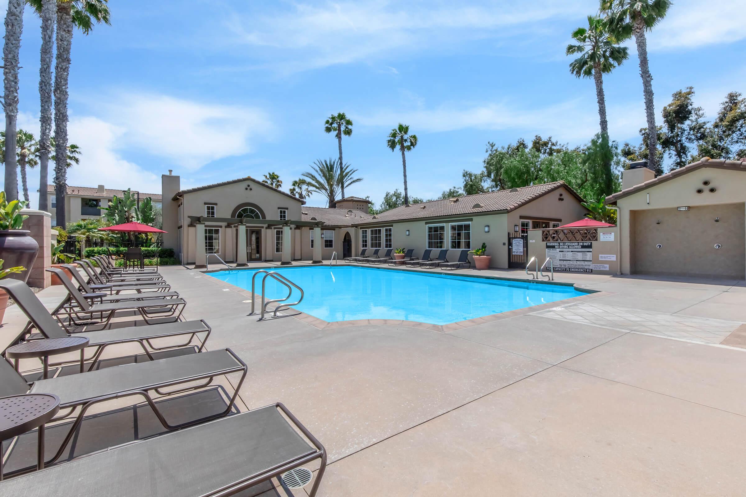 A sunny, outdoor pool area featuring a clear blue pool surrounded by lounge chairs. Palm trees line the background, and there are shaded seating areas with umbrellas. Buildings with a Mediterranean style are visible nearby, along with a clear sky and a well-maintained patio area.