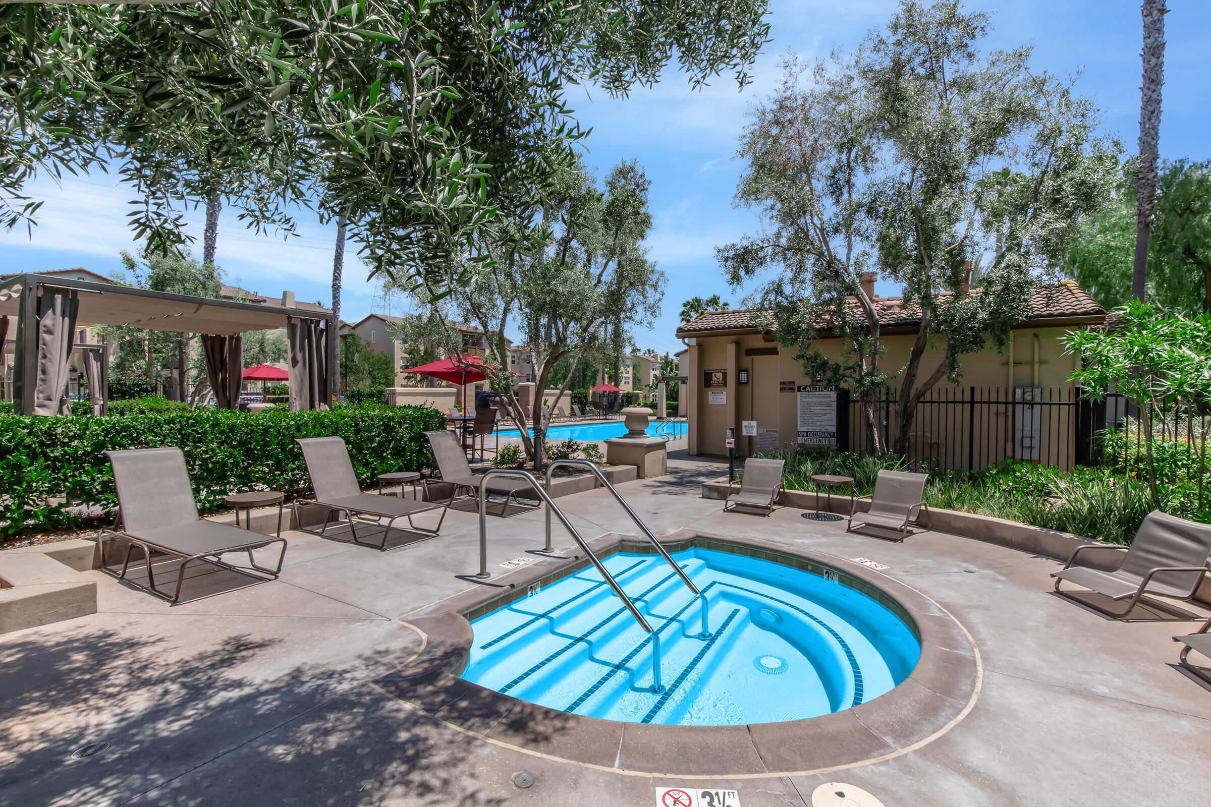 A scenic outdoor area featuring a hot tub surrounded by lounge chairs, with lush greenery and trees in the background. A pool can be seen nearby, along with shaded seating and umbrella coverings. The atmosphere appears inviting and relaxing, ideal for leisure and enjoyment.