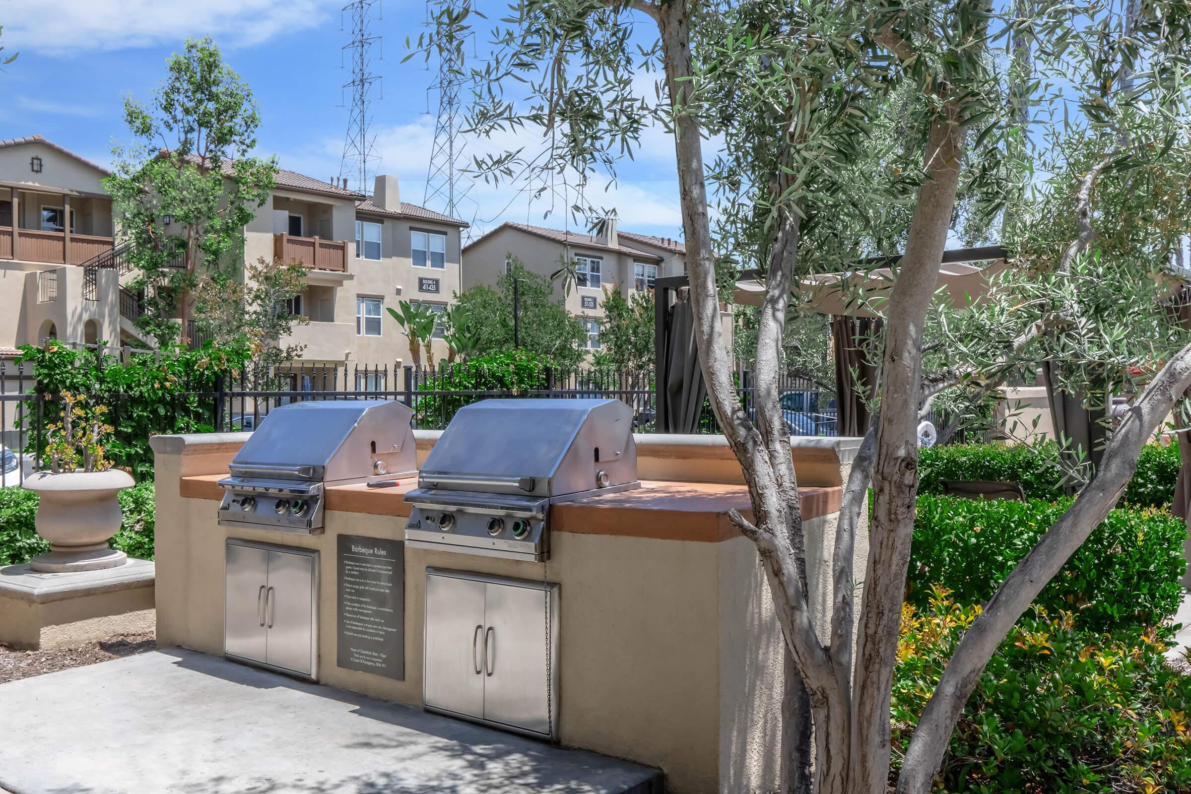 Outdoor grilling area featuring two stainless steel barbecue grills with a stone countertop surrounded by greenery. In the background, a residential complex is visible along with various trees and shrubs, providing a pleasant setting for outdoor cooking and entertaining.