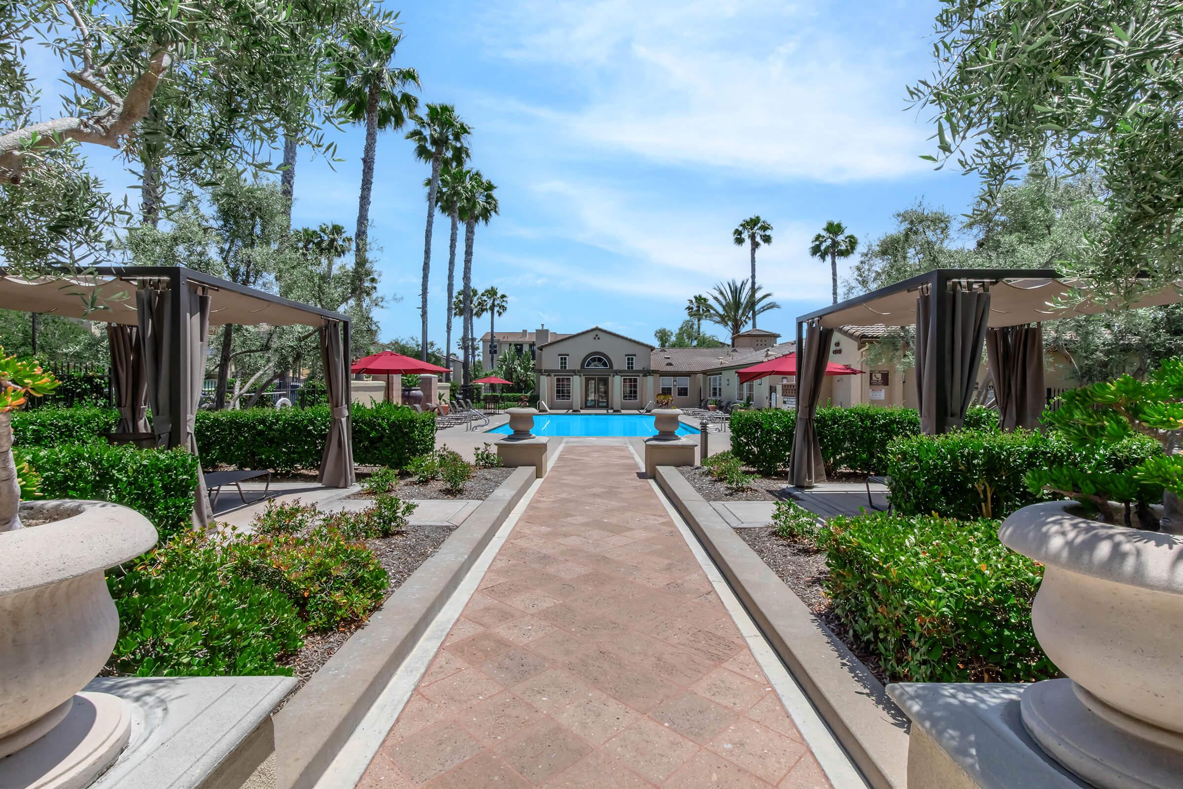 A landscaped garden area featuring a central pool, flanked by cabanas and surrounded by decorative bushes and palm trees. The pathway leading to the pool is lined with planters, and red umbrellas provide shaded seating. The background includes a stylish building with an arched entrance. Clear blue skies are visible above.