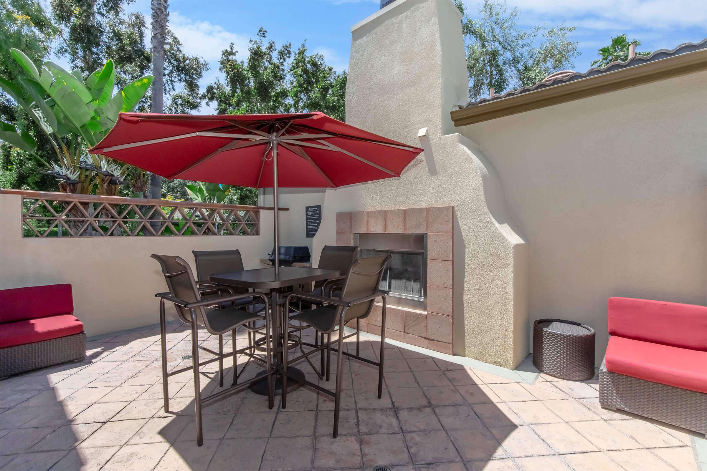 An outdoor seating area featuring a rectangular table and chairs under a red umbrella, with a stone fireplace in the background. Two red lounge chairs are placed nearby, surrounded by greenery and trees, creating a cozy and inviting atmosphere. Bright sky with scattered clouds visible above.