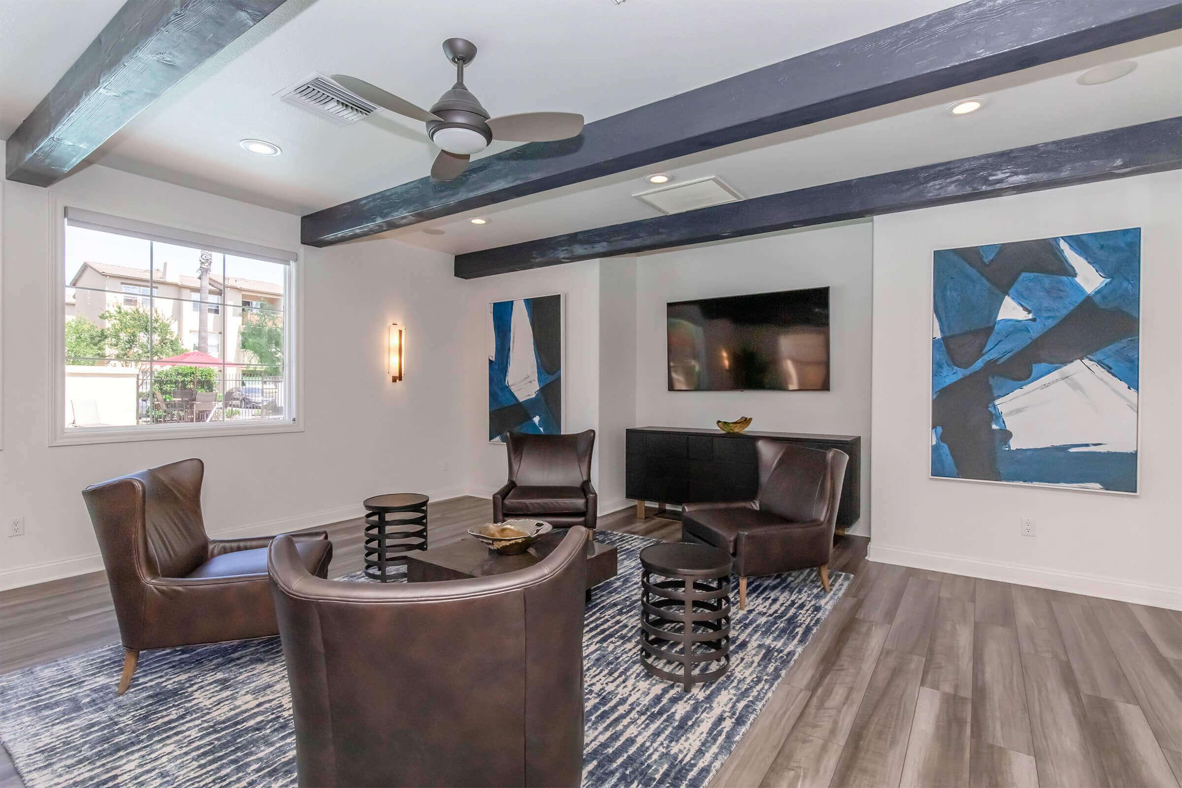 A modern living room featuring two dark leather chairs, a sleek TV mounted on the wall, and a large abstract painting. Sunlight filters through a window, illuminating the space which has gray wood flooring and black wooden beams on the ceiling. A stylish area rug adds texture to the setting.
