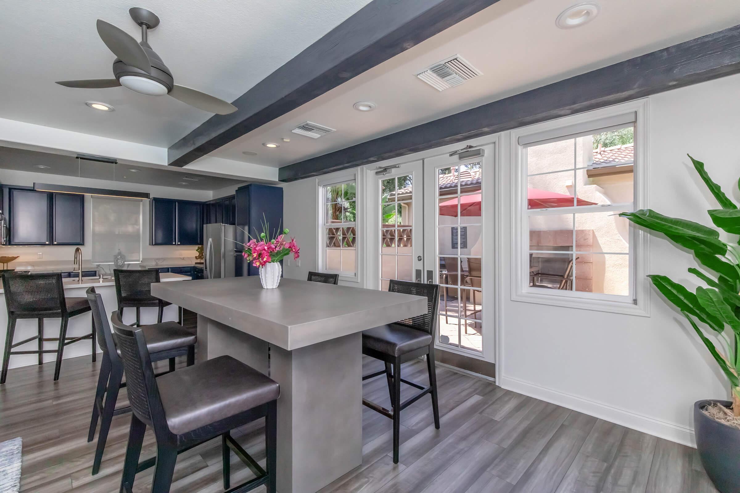 Modern kitchen and dining area featuring a spacious gray countertop island with dark bar stools, blue cabinetry, and stainless steel appliances. Large windows invite natural light, revealing a patio outside with a red umbrella. A potted plant adds a touch of greenery to the stylish interior.