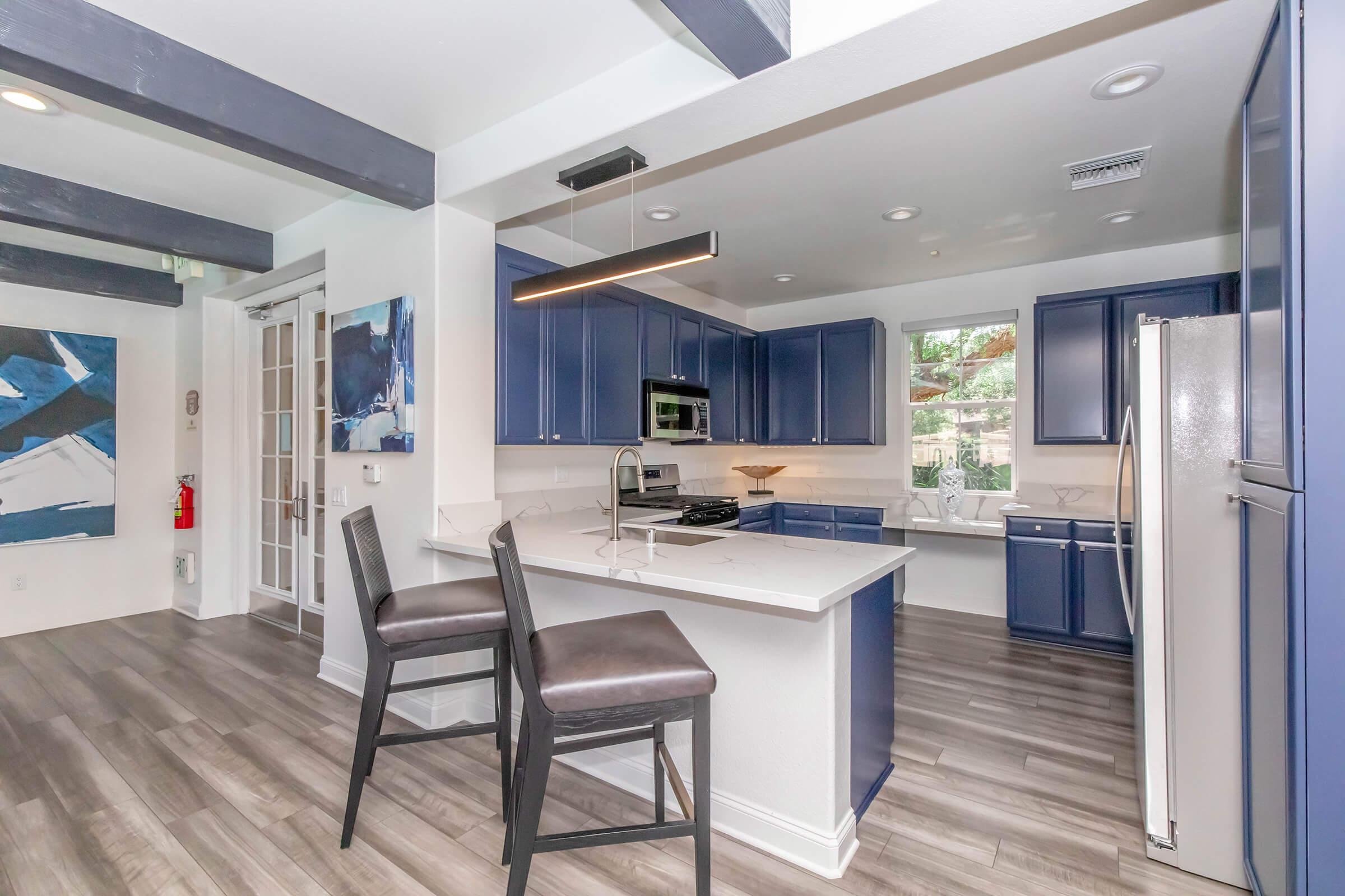 Modern kitchen featuring blue cabinetry, stainless steel appliances, and a spacious island with seating. The room has a light and airy feel, with large windows allowing natural light to flow in. Decorative beams line the ceiling, and contemporary artwork is displayed on the wall.