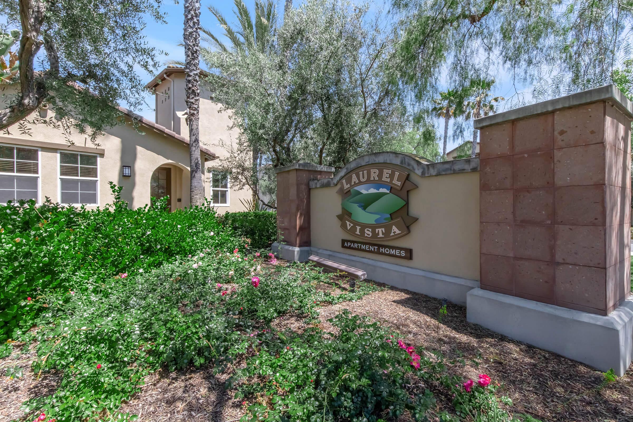 Sign for Laurel Vista Apartment Homes, surrounded by greenery and pink flowers. The entrance features a stone structure with lush landscaping, palm trees, and a sunny sky in the background. The design reflects a welcoming atmosphere for residents and visitors.