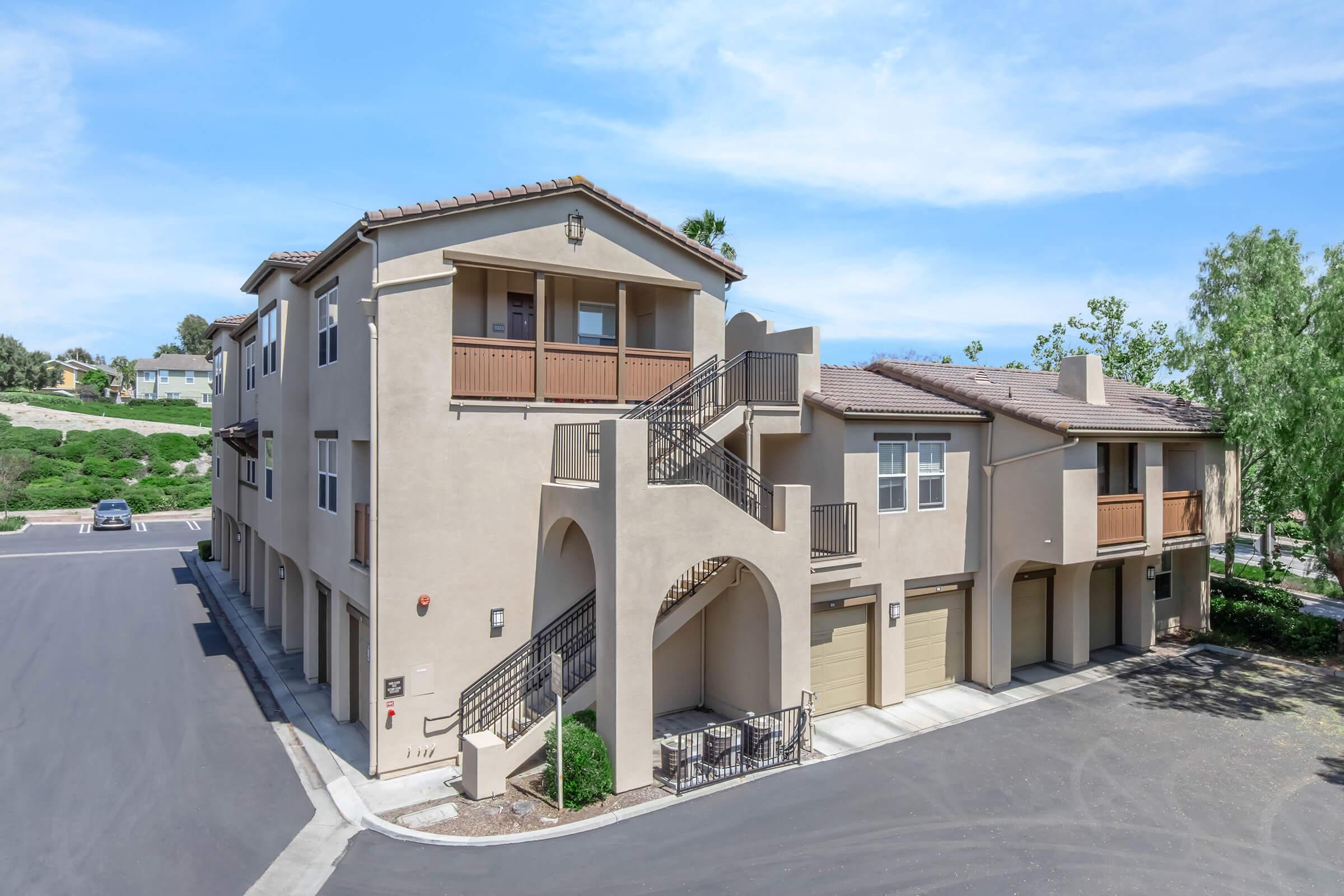 A multi-story residential building with a Mediterranean architectural style. It features a staircase leading to the second floor, balconies with wooden railings, and garage doors on the ground level. The surrounding area includes neatly landscaped greenery and a clear blue sky.