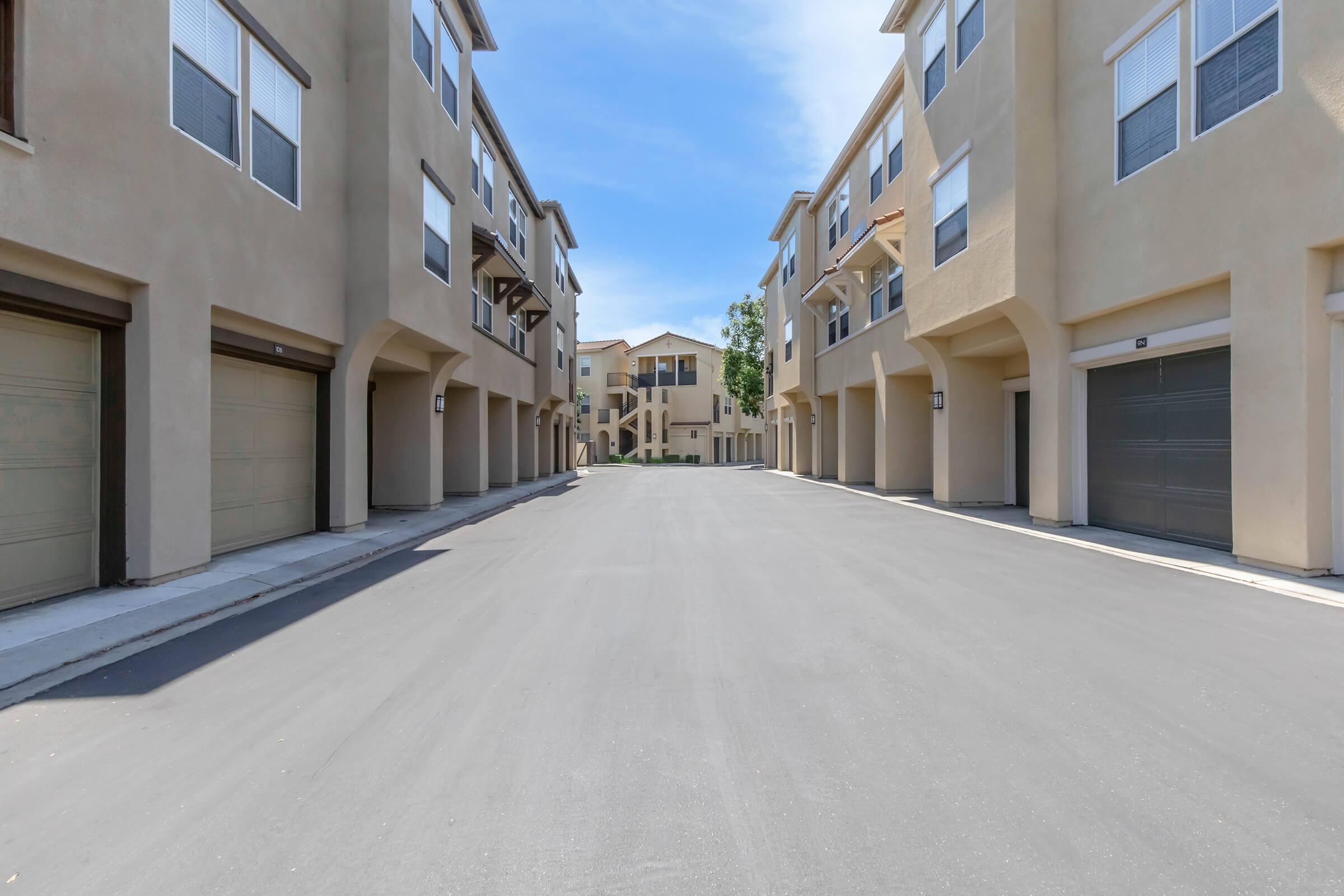 A wide view of a residential area featuring two rows of modern, multi-story buildings with garages. The street is paved and empty, with blue skies and clouds visible in the background. The architecture is contemporary, with multiple windows on each building. Trees are planted along the sides of the street.