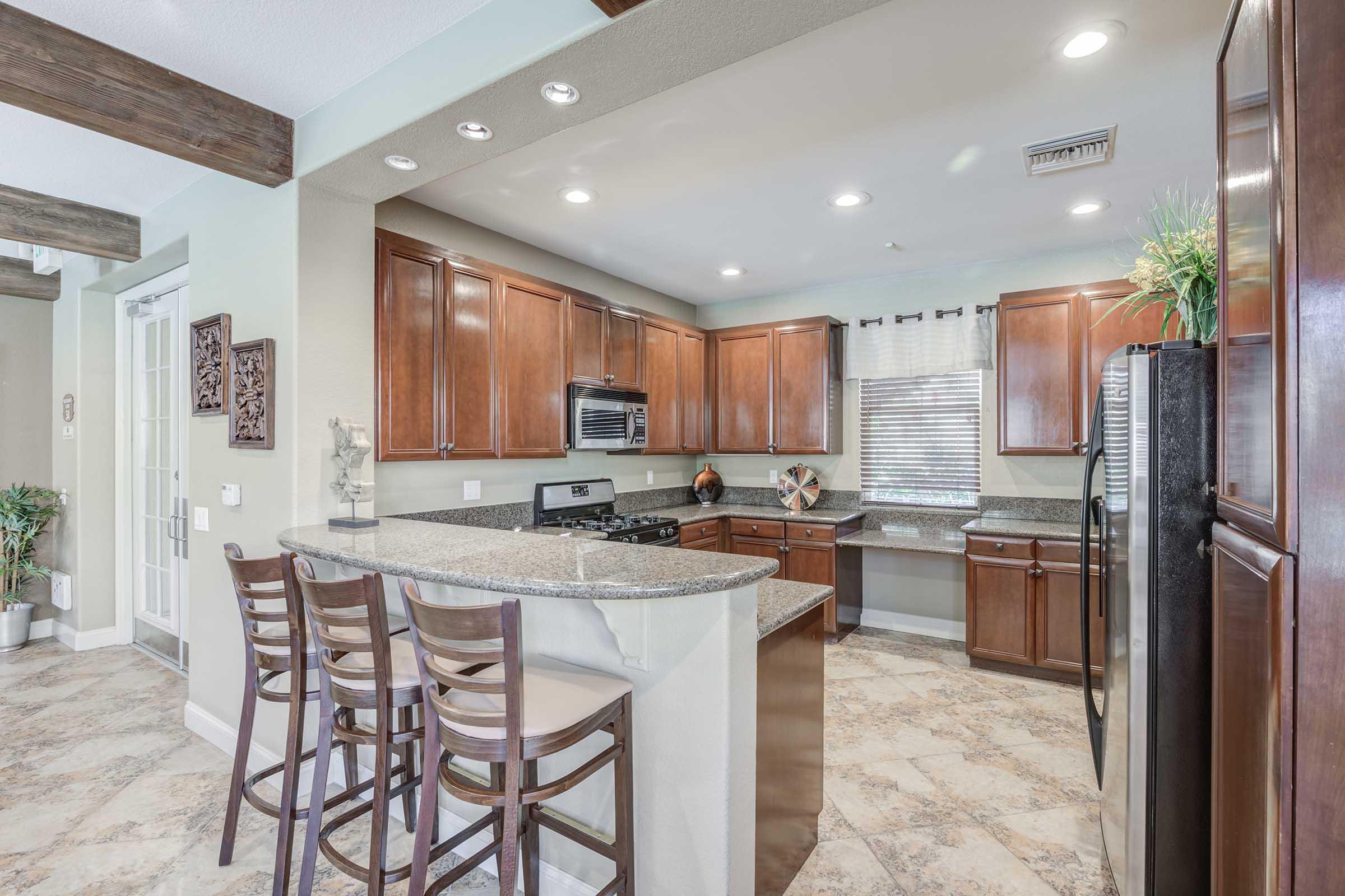 Modern kitchen featuring wooden cabinets, granite countertops, and a breakfast bar with four stools. The room includes stainless steel appliances, large windows, and tile flooring.