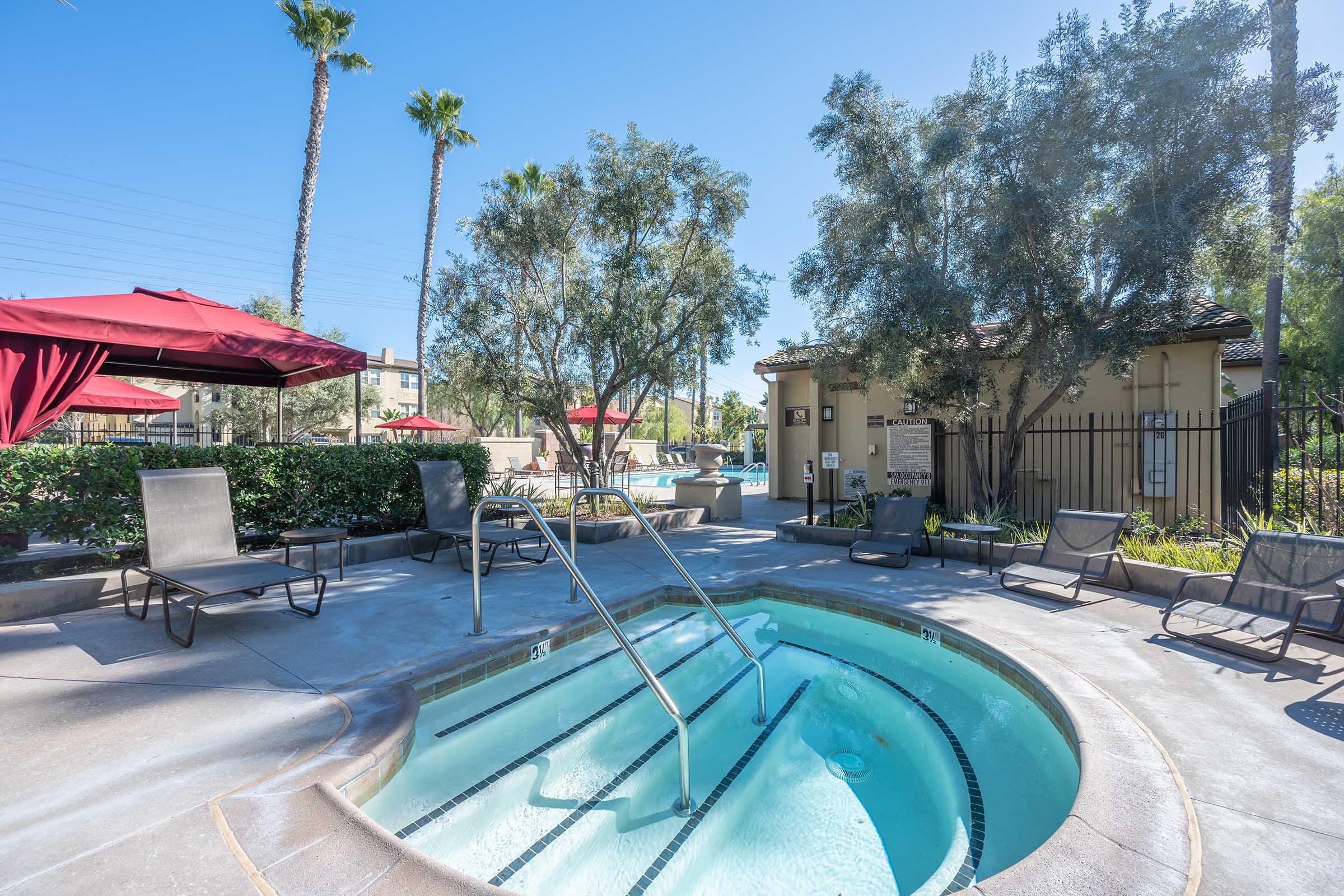 A clear blue hot tub surrounded by lounge chairs, palm trees, and red umbrellas in a sunny pool area.