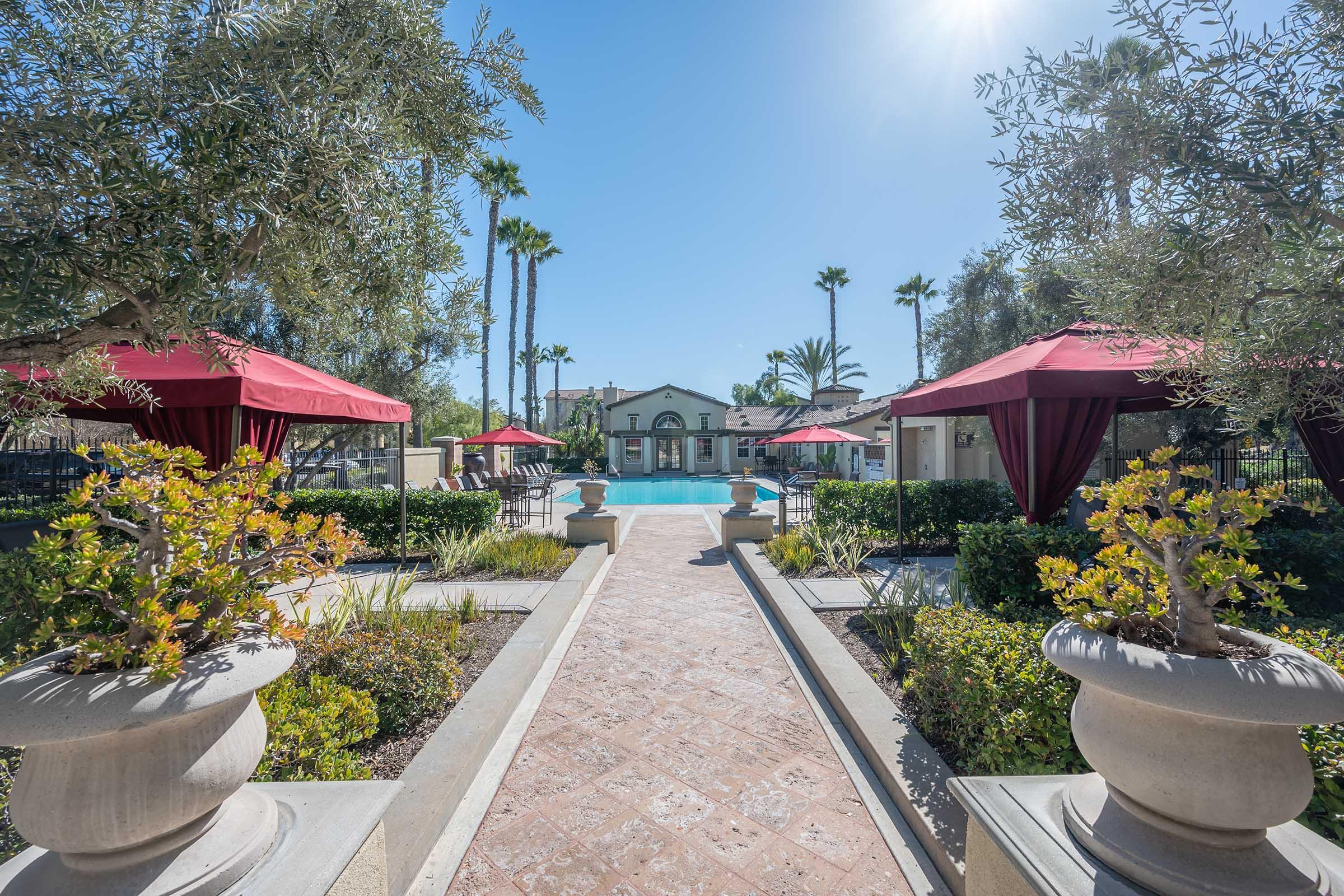 View of a serene outdoor pool surrounded by lush greenery and palm trees, with red canopies providing shade. Elegant urns with plants line the pathway leading to the pool area, creating a tranquil atmosphere.