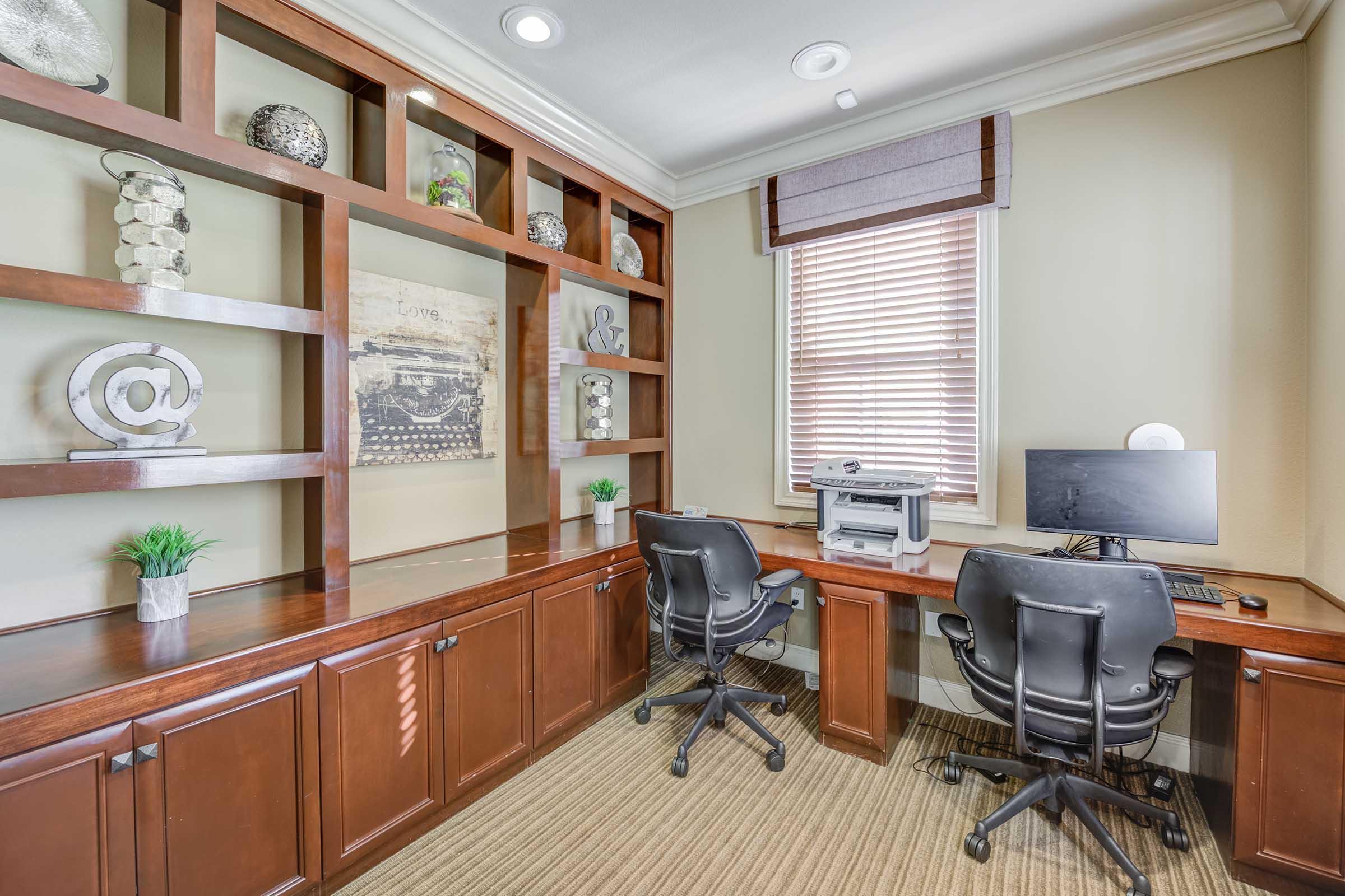 A home office featuring two black chairs at a wooden desk, a computer monitor, and a printer. The shelves above the desk are decorated with various decorative items and plants, and there is a window with blinds on the right side. The walls are painted a neutral color, creating a modern and organized atmosphere.