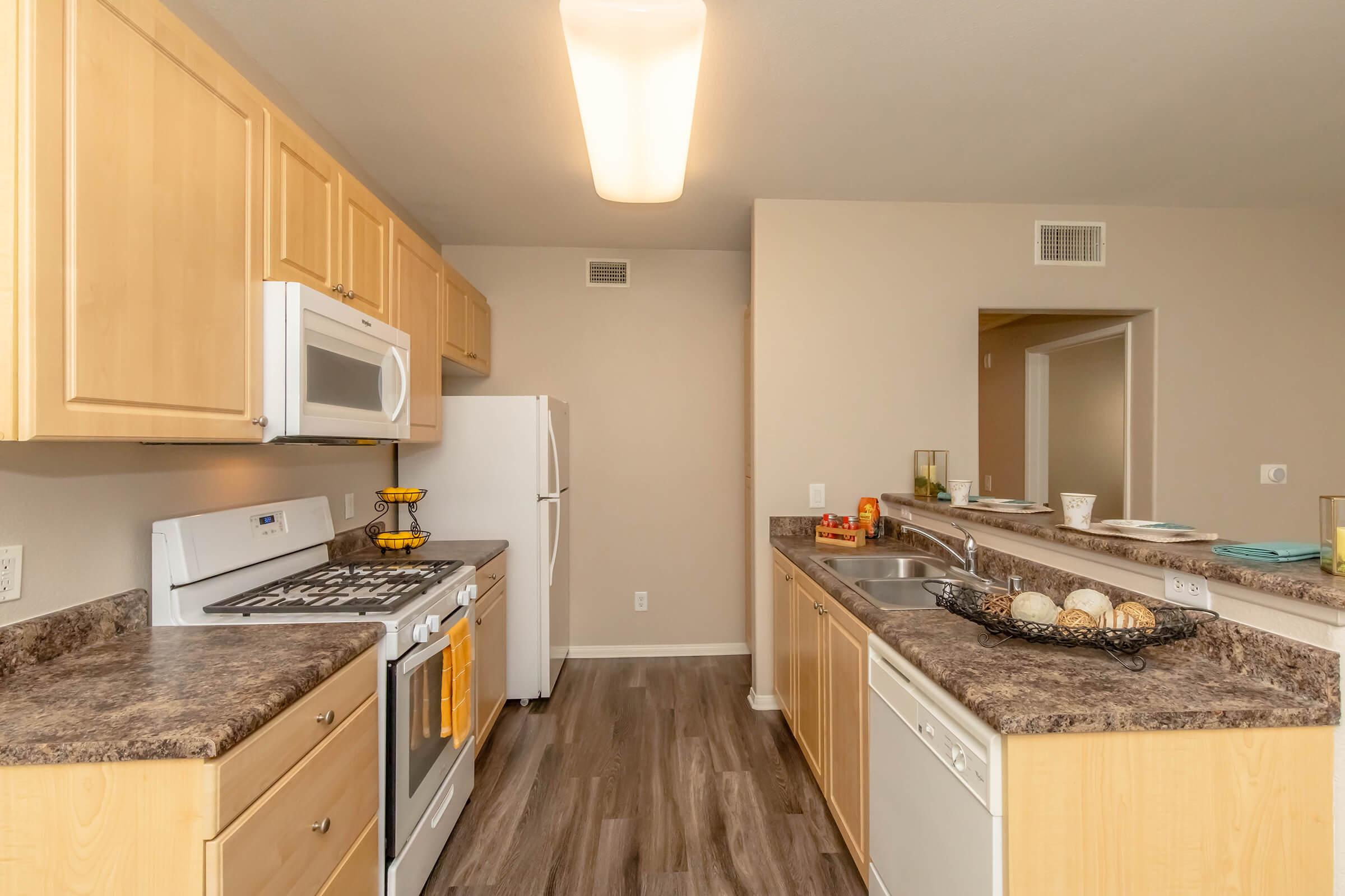 A modern kitchen featuring light wood cabinetry, a gas stove, microwave, and white refrigerator. The countertop is dark with a sink and dishwasher. Natural light comes from a ceiling fixture, and the flooring is a wood-like material, creating a warm and inviting atmosphere.
