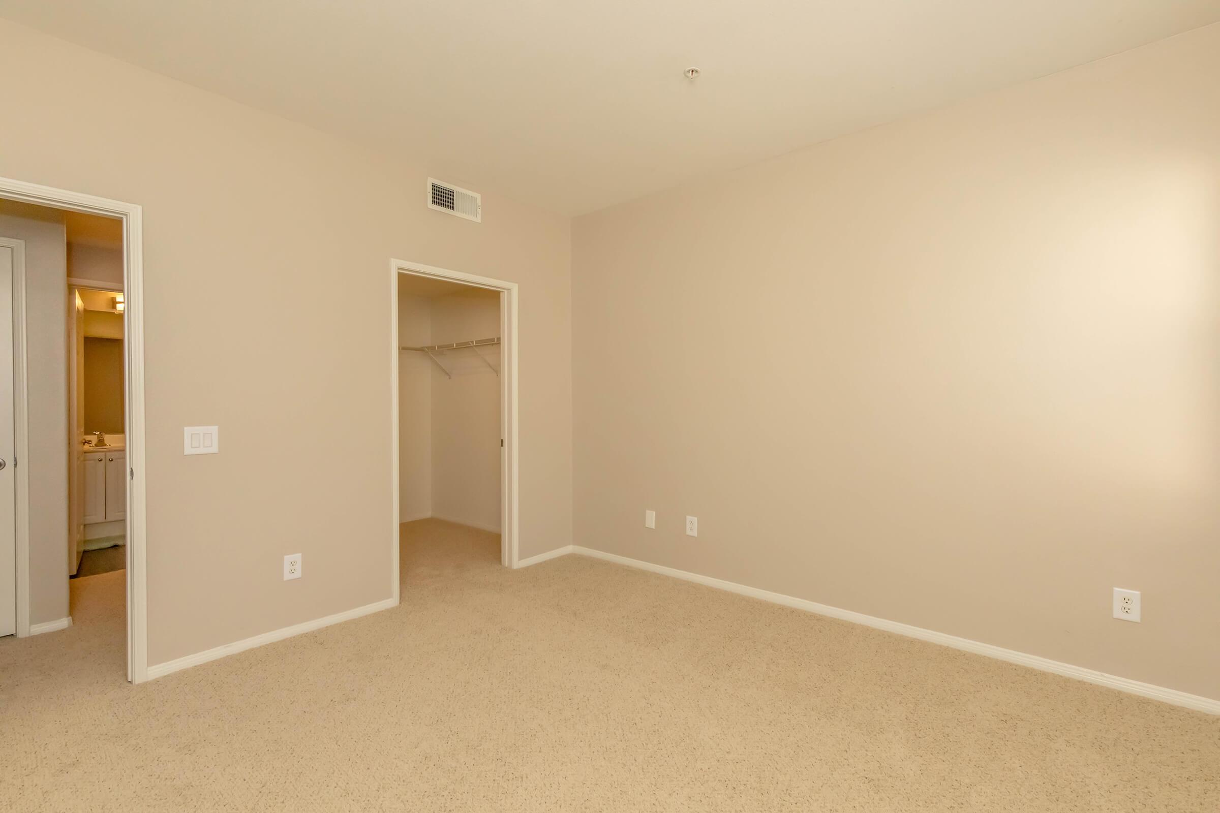 A spacious, empty bedroom with light beige walls and carpet. It features a closet with an open door, white light switches on the wall, and a doorway leading to a bathroom area visible in the background. The room is well-lit and has a minimalistic, tidy appearance.