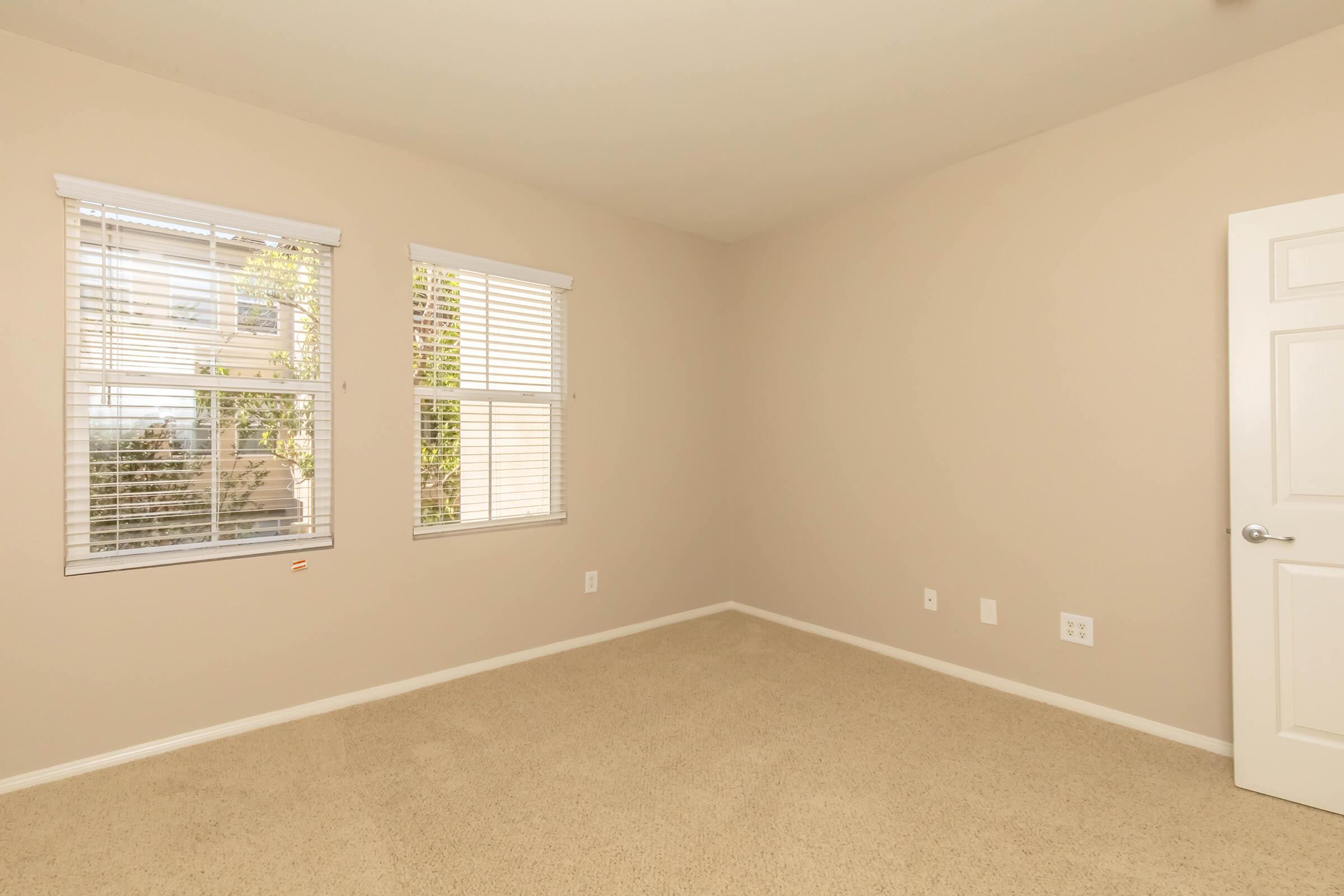 Interior of a vacant room with beige walls and carpet, featuring two windows with blinds that allow natural light. The room is empty, with a plain white door on one side and simple electrical outlets and light switches visible on the walls.