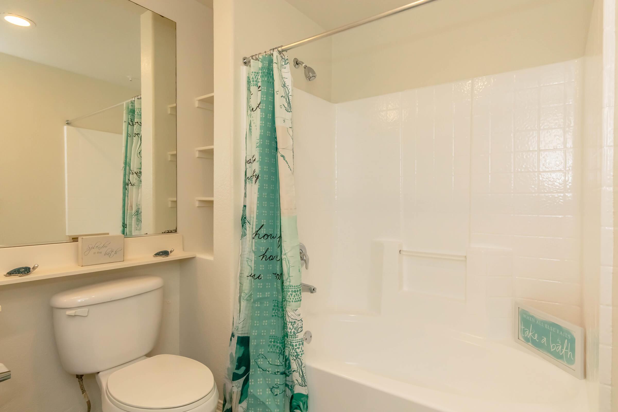 A clean bathroom featuring a white bathtub, toilet, and a mirror above a countertop. The shower curtain displays a blue and green map design, while decorative signs on the countertop encourage relaxation. Bright lighting enhances the overall fresh and inviting atmosphere.
