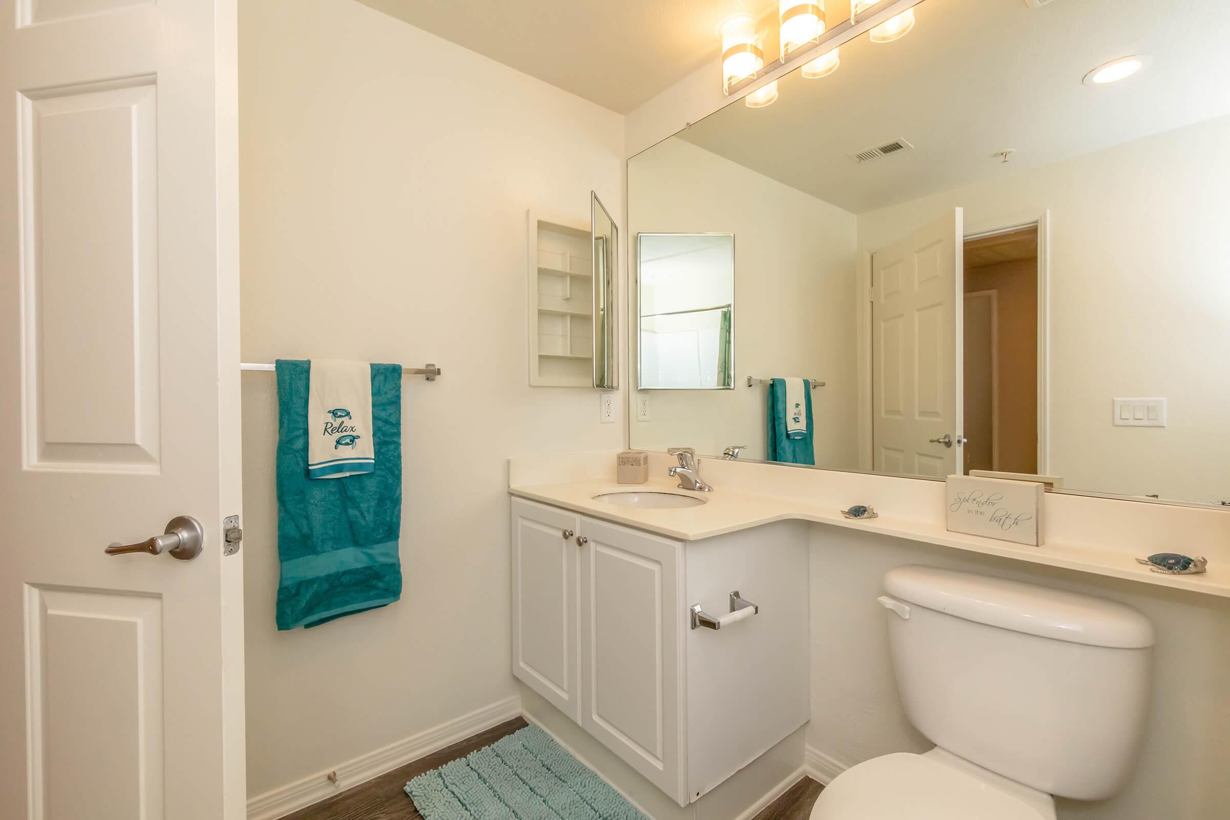 A clean and modern bathroom featuring a white toilet, a sink with a mirrored cabinet above, a light-colored countertop, and towels in teal. The walls are painted in a light shade, and there is a small decorative sign on the counter. The flooring is wooden laminate, adding a warm touch to the space.