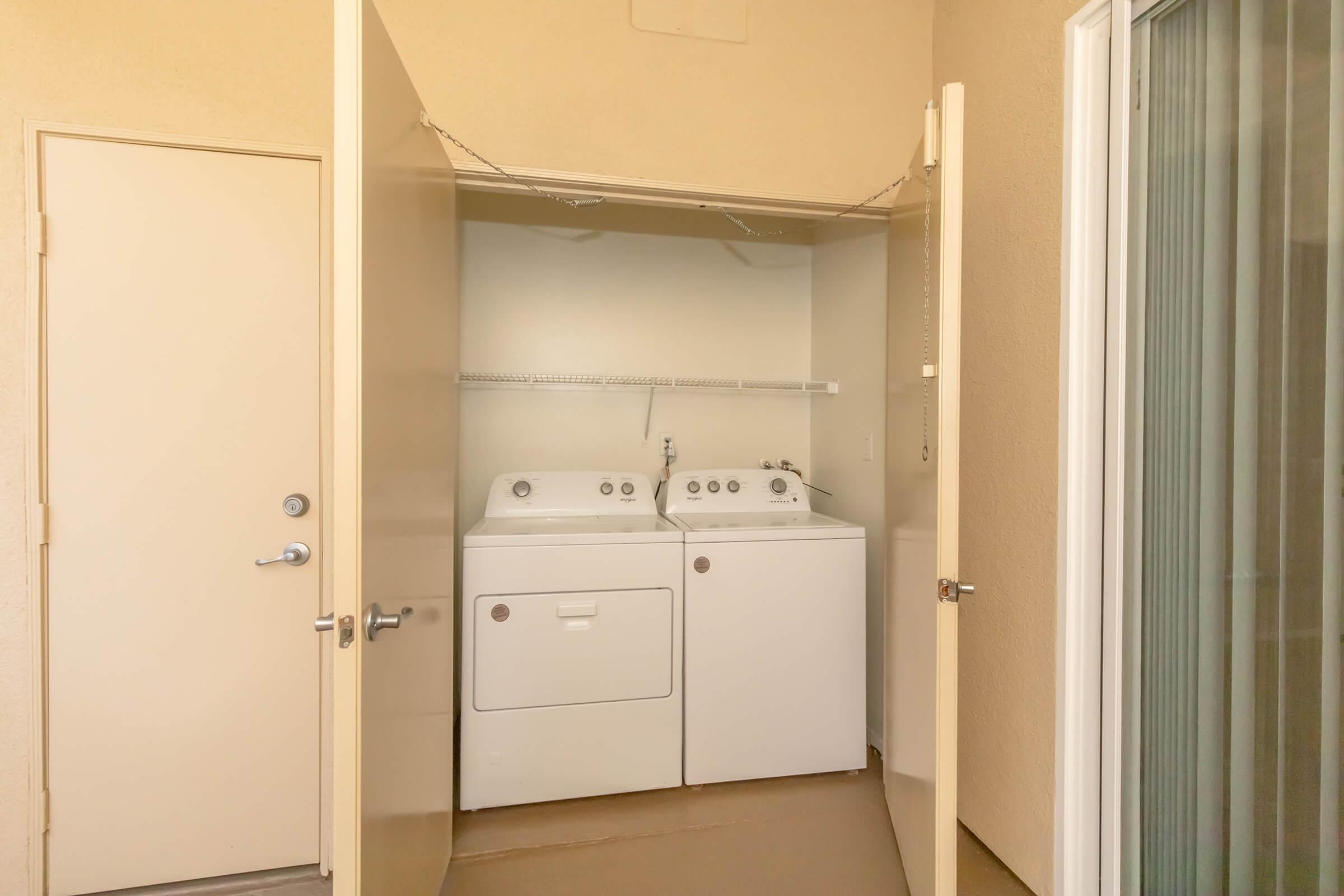 A laundry closet with a stacked washer and dryer, featuring white appliances and a shelf above. The closet doors are open, revealing the machines, and there is a door leading to an outside area. The walls are a neutral color, and there is a sliding glass door on the right.