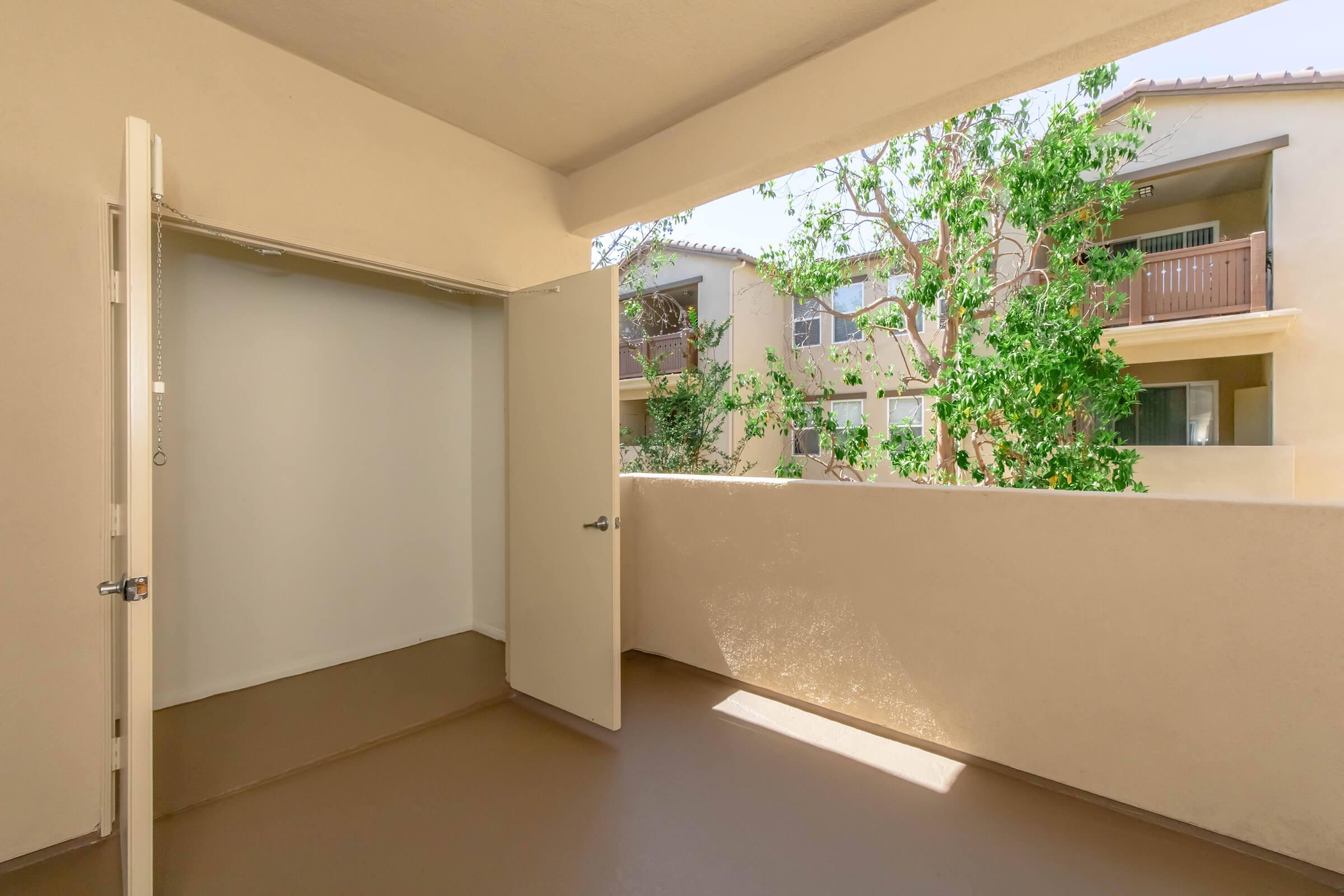 View of a balcony with an open door leading to a storage area. The balcony features a beige wall and railings, with greenery visible from nearby trees and neighboring buildings. The area has a clean and airy atmosphere.