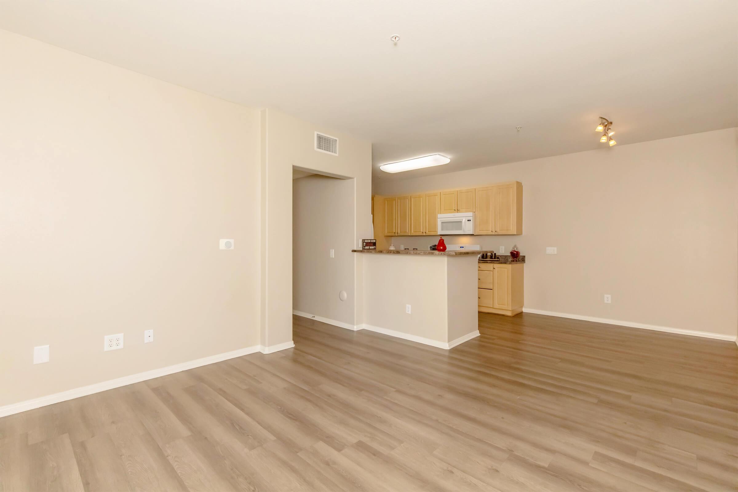 A spacious, empty apartment interior featuring light-colored walls and wood-like flooring. There is a kitchen area with wooden cabinets and a white microwave, and an open layout that connects the living area to the kitchen. The space is well-lit and unadorned, creating a modern, minimalist feel.
