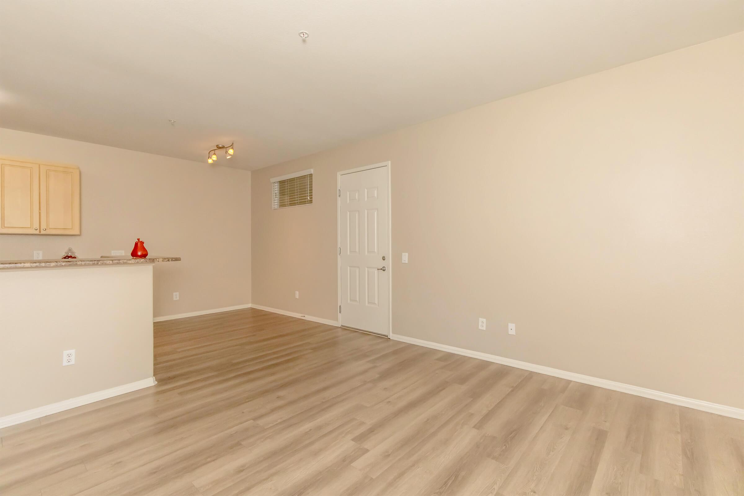 A spacious, well-lit living area with light beige walls and wooden flooring. There's a small kitchen area to the left with wooden cabinets, and a white door leading to another room. The space is empty, creating a clean and open feel.