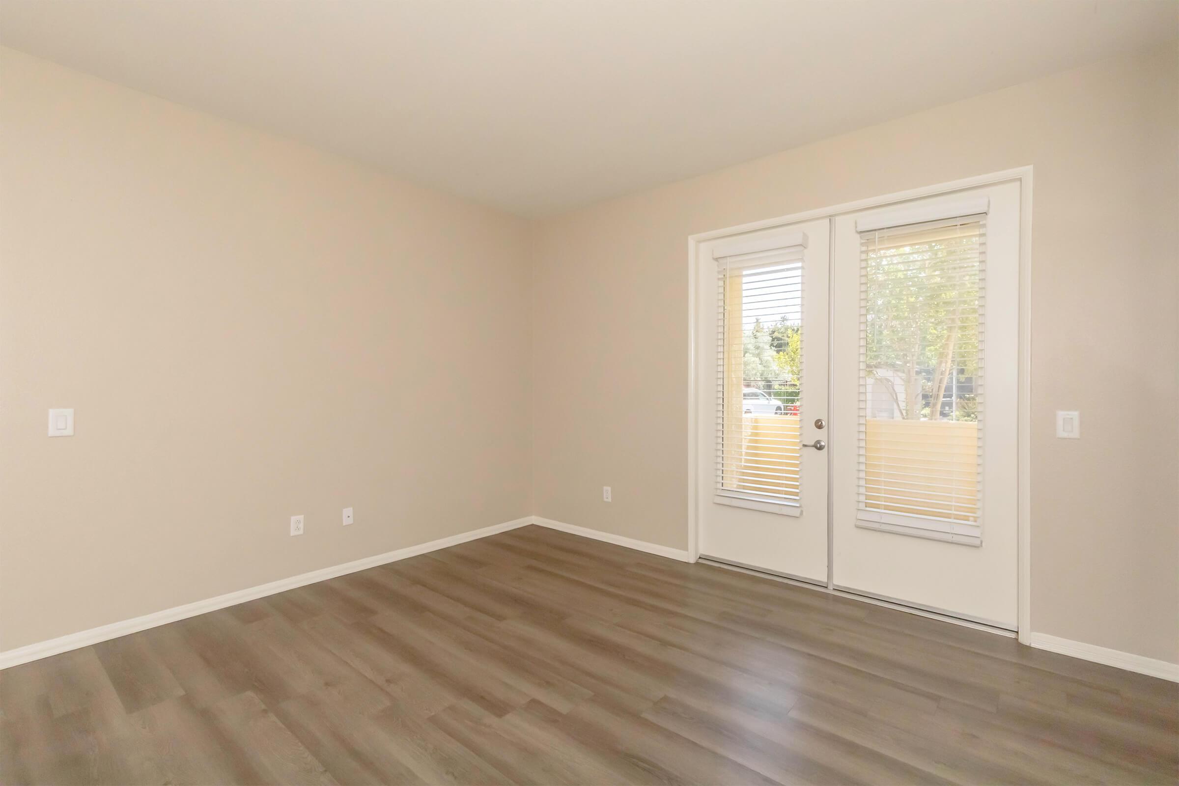 A spacious, empty room with light-colored walls and hardwood flooring. Large glass doors with vertical blinds lead to an outdoor area, allowing natural light to enter. The room is unfurnished, providing a blank canvas for decoration and personalization.