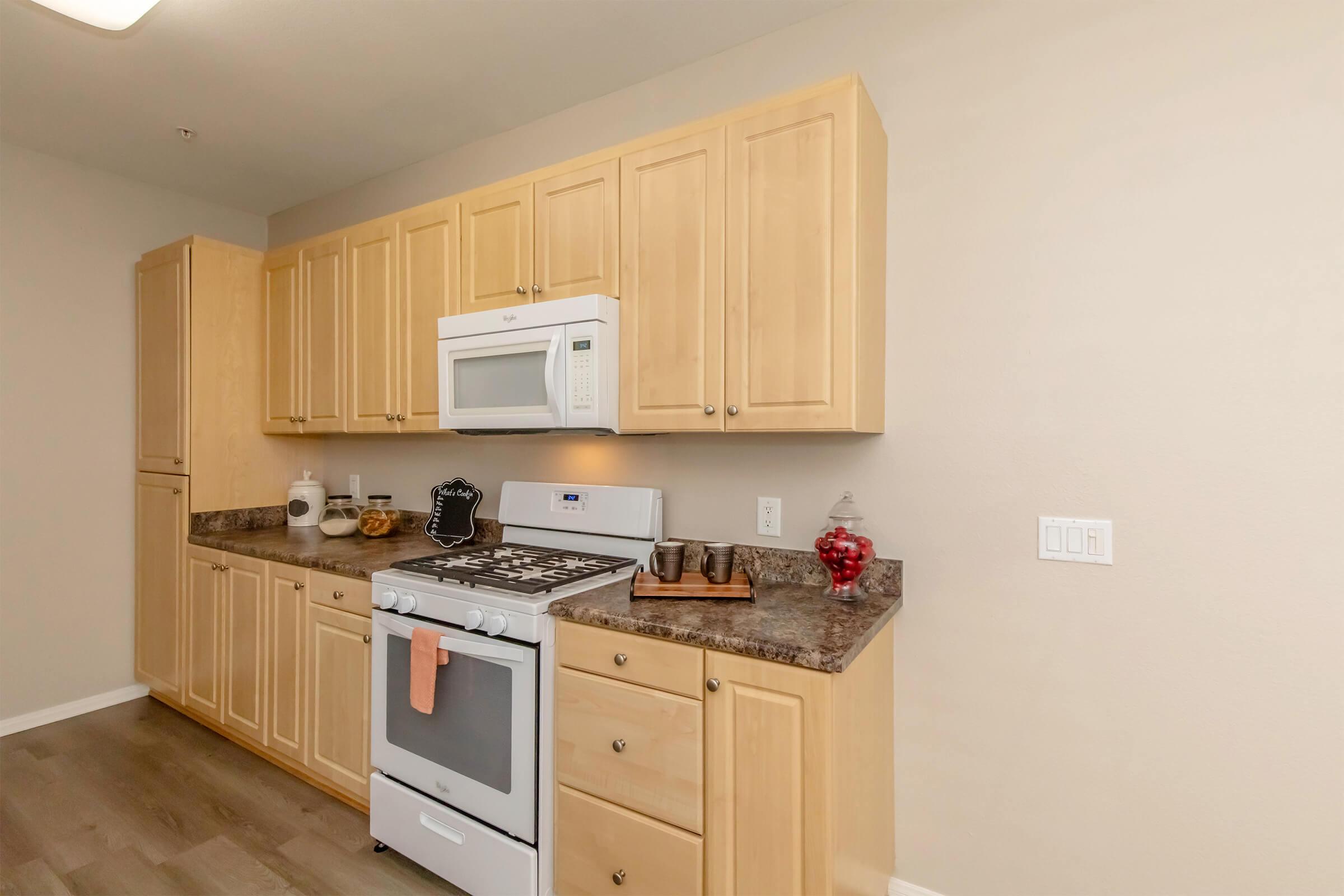 Modern kitchen with light wood cabinetry, a white gas stove with an oven, a built-in microwave, and granite countertops. A few kitchen accessories and a jar of red candy are displayed on the counter. The walls are a neutral color, and the flooring is light wood.